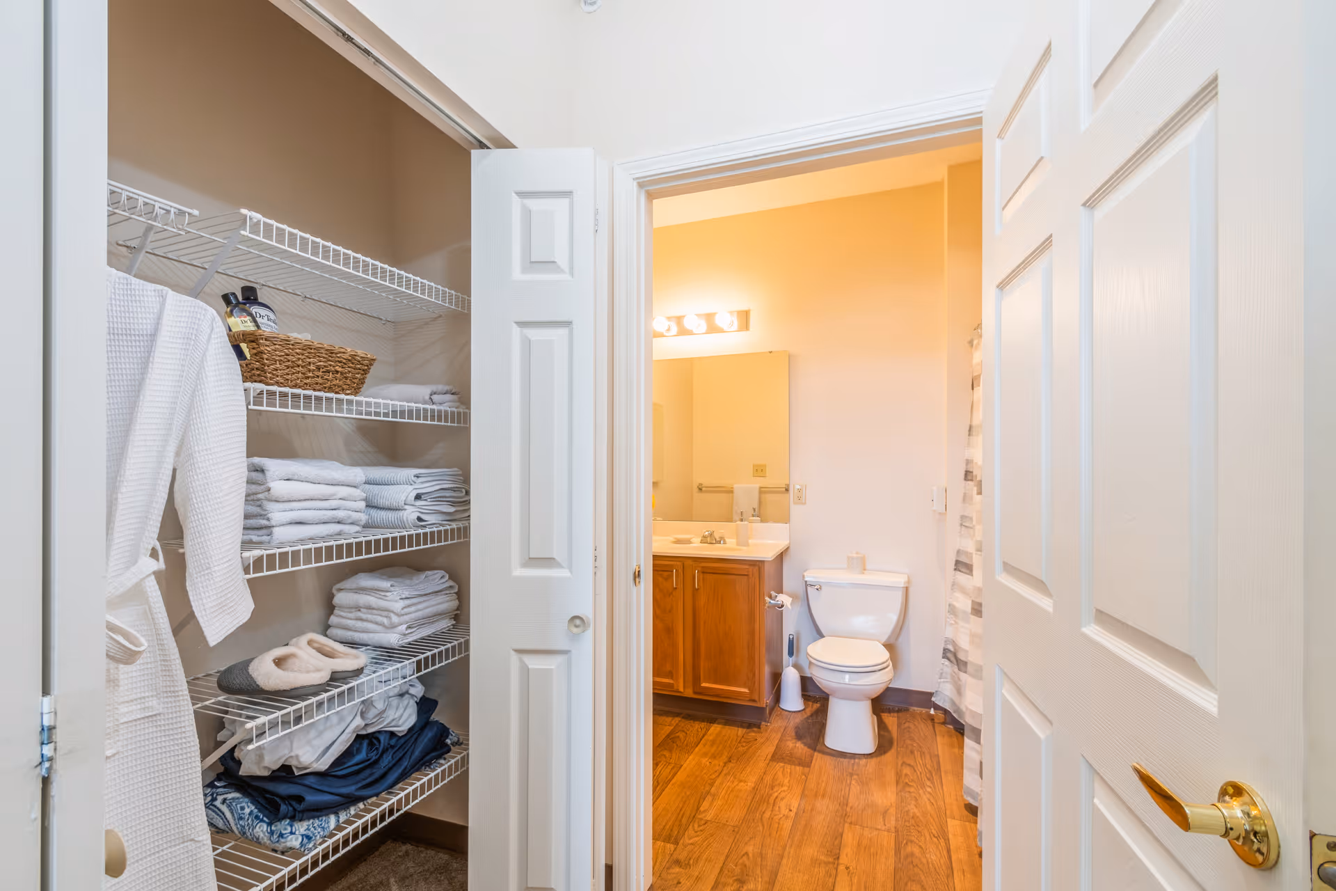 View of a bathroom with a wooden floor, a white toilet, a wooden vanity with a sink and mirror, and a shower curtain. To the left, there is an open closet with wire shelves holding folded towels, a basket, slippers, and a hanging white robe.
