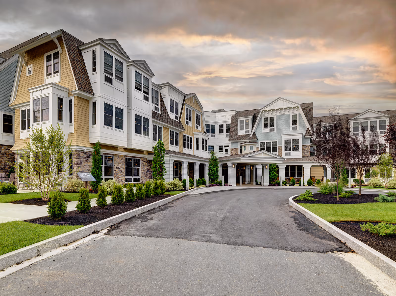 Exterior view of a large, multi-story senior living facility with a driveway leading to a covered entrance. The building features a mix of stone and siding with multiple windows and landscaped greenery around the entrance. The sky is partly cloudy with a warm sunset glow.