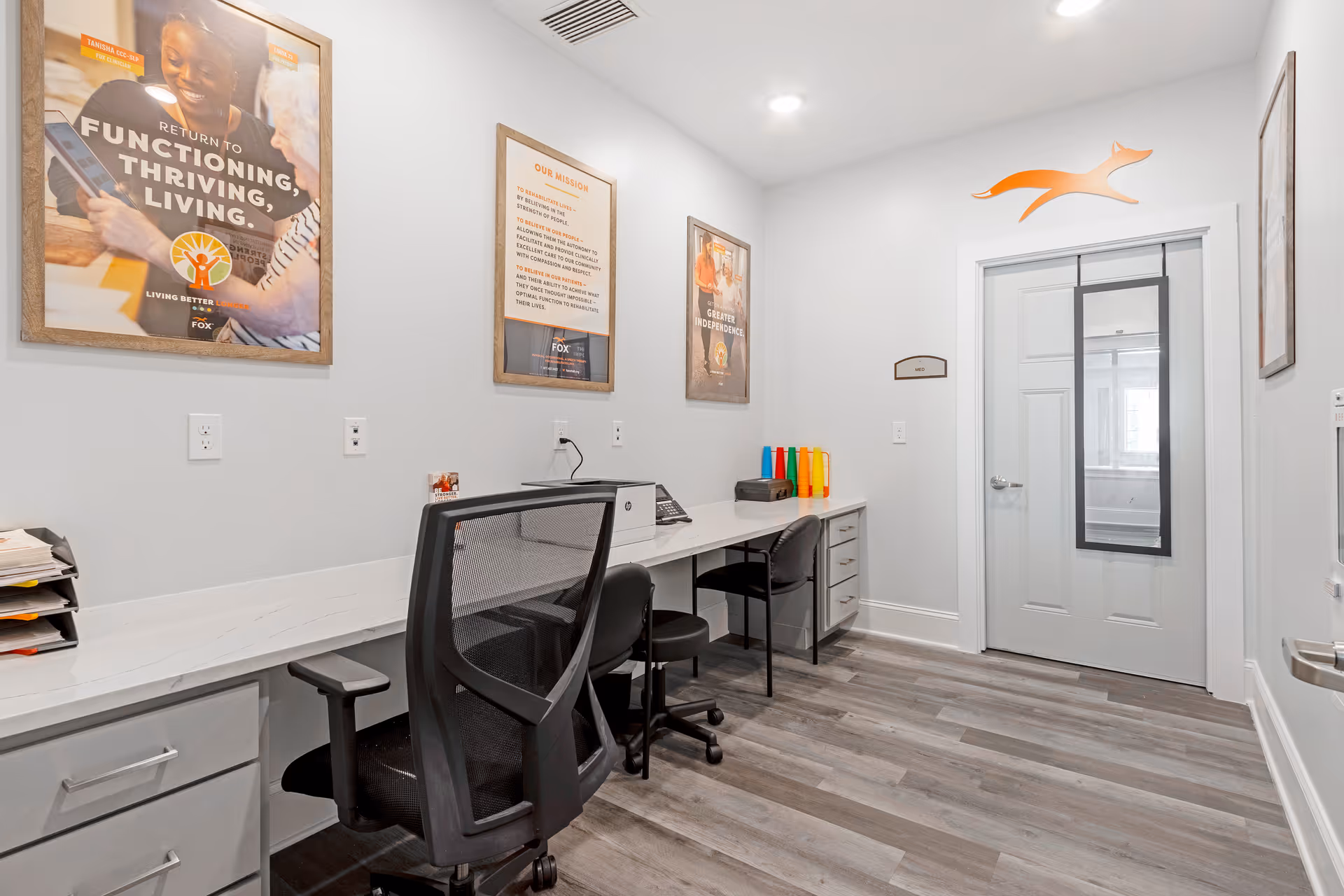 A small office or work area with a long white desk against a light gray wall. There are three black chairs, two of which are office chairs, and a few colorful cones on the desk. The walls have three framed posters with motivational and mission statements. The floor is covered with gray wood-like vinyl planks. A white door with a window and an orange fox logo above it is visible at the end of the room.