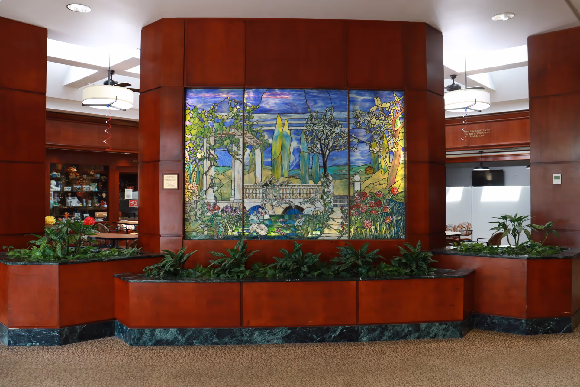 Interior view of a senior living facility featuring a large stained glass artwork depicting a garden scene with trees, flowers, and a bridge. The artwork is set in a wooden panel wall with built-in planters containing green plants at the base. The space has carpeted flooring and ceiling lights, with glimpses of seating areas and a small shop in the background.