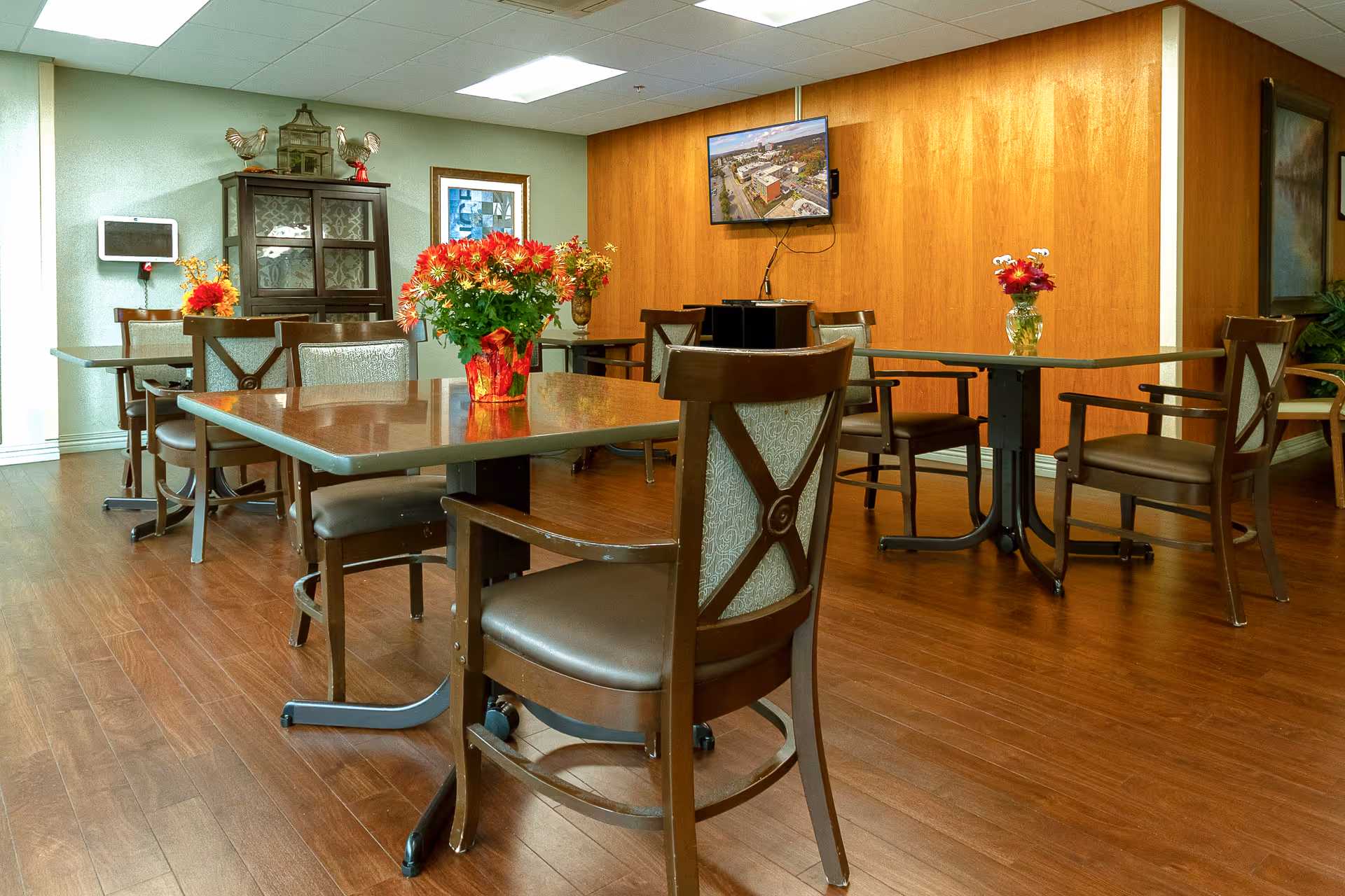 A dining area with several tables and chairs arranged on a wooden floor. Each table has a vase with colorful flowers. There is a wooden cabinet against the wall, a wall-mounted TV displaying an aerial view of a building, and a framed picture on the wall. The room has a warm and inviting atmosphere.