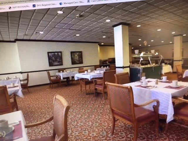 A dining room in a senior living facility with several tables covered in white tablecloths, set with cups and plates. The room has patterned carpet flooring, wooden chairs with armrests, and framed artwork on the walls. There is a buffet area in the background with food warmers and serving utensils.