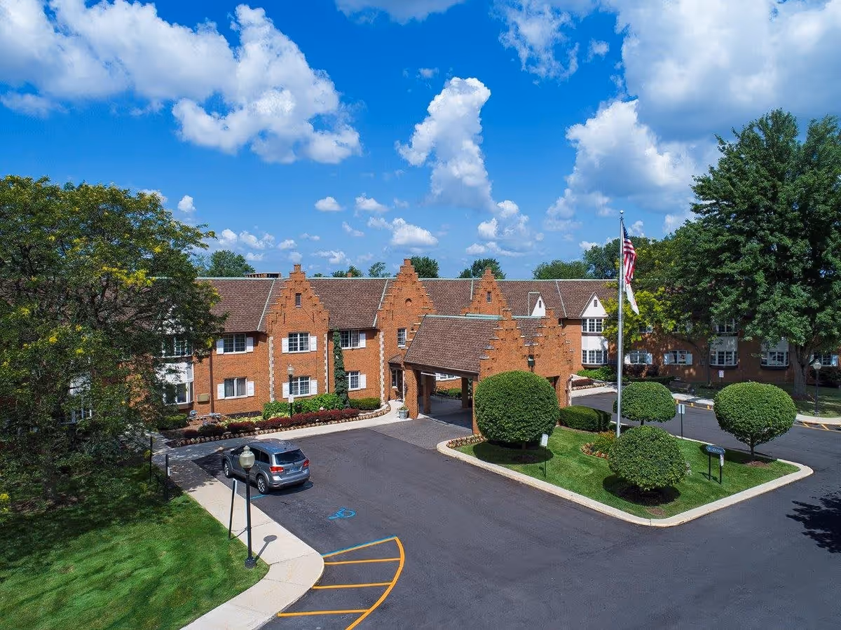 Exterior view of a senior living facility named American House East I featuring a large brick building with a peaked roof entrance, neatly trimmed bushes, an American flag on a flagpole, a parked car, and a bright blue sky with scattered clouds.
