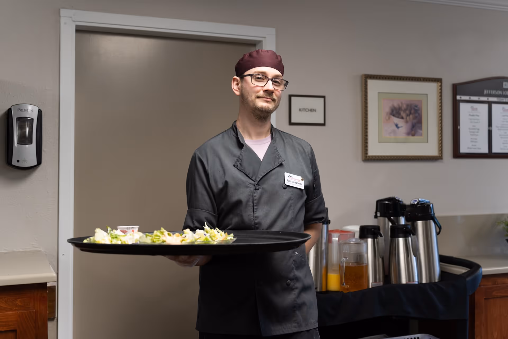 A man wearing a dark chef uniform and a maroon cap is holding a large tray with plates of salad. He is standing indoors in front of a beige wall with framed pictures and a sign that reads 'KITCHEN'. There is a beverage station with coffee dispensers and juice pitchers behind him.