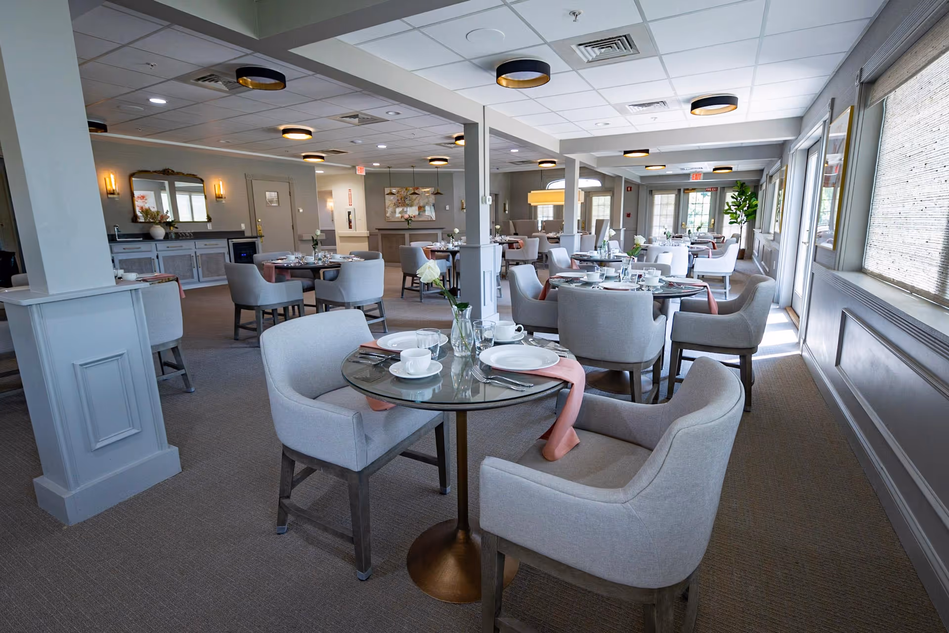 A spacious and well-lit dining room with multiple round tables set with white plates, cups, and silverware. Each table has comfortable upholstered chairs and a small vase with a flower. The room features neutral tones, large windows with blinds, ceiling lights, and decorative wall sconces, creating an inviting atmosphere.
