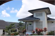 Exterior view of a nursing and rehab center building with a tiled roof, white walls, glass block windows, and surrounding tropical plants under a partly cloudy sky.