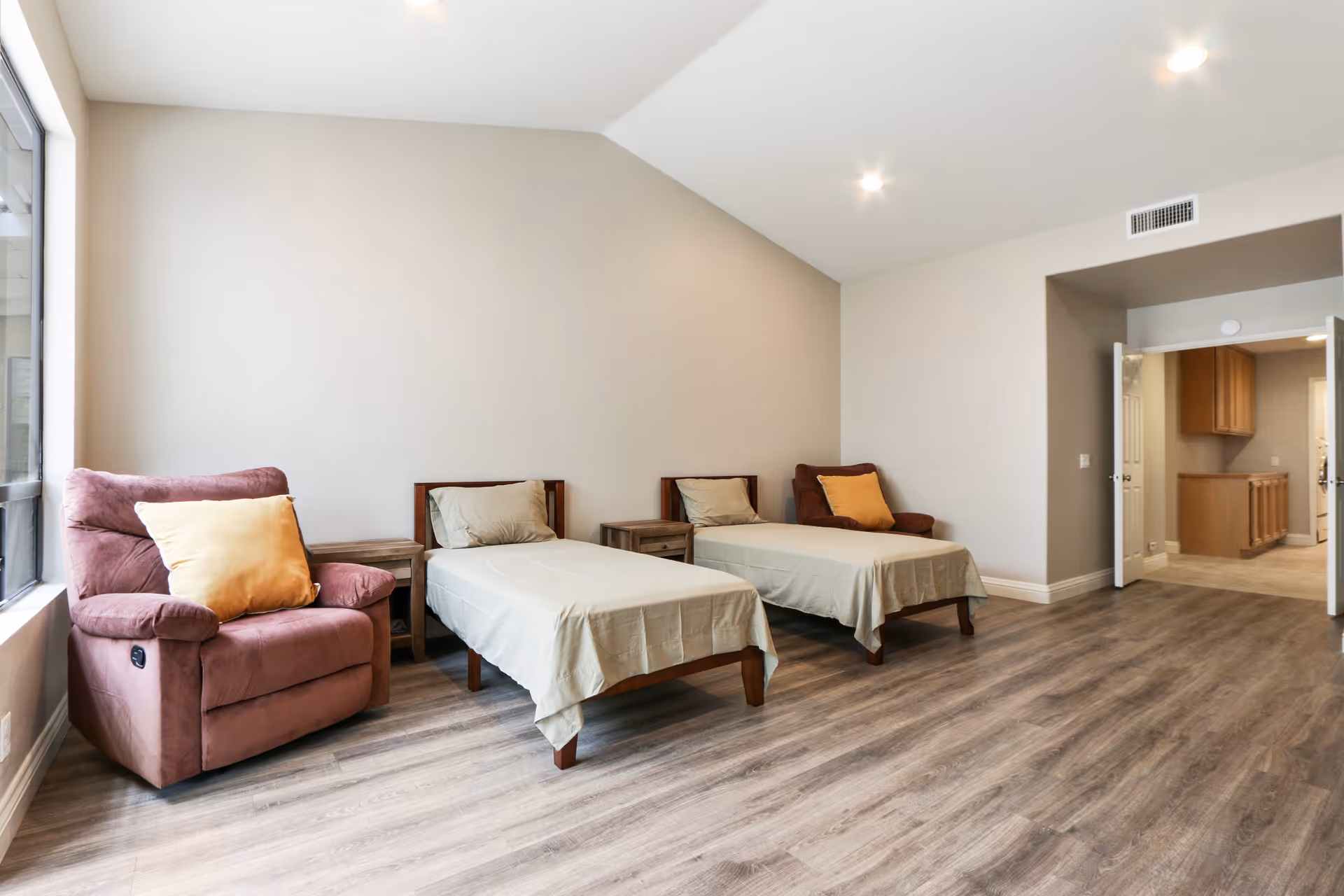 Shared bedroom with two single beds, a pink recliner with a yellow pillow, bedside tables and wood-look flooring.