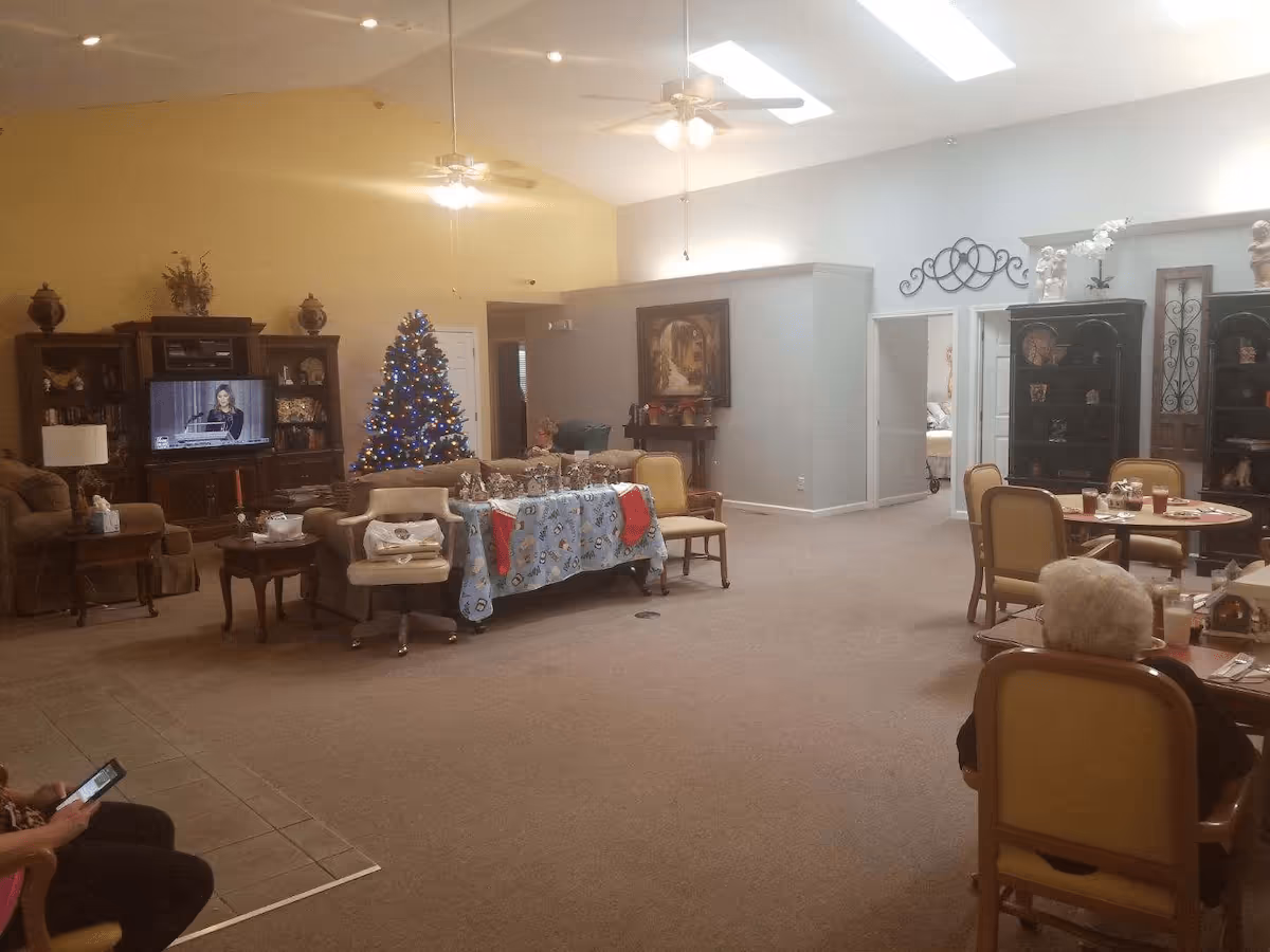 A spacious living room area in an assisted living community decorated for Christmas with a Christmas tree, a table covered with a festive cloth and stockings, several chairs and sofas, a television on a wooden cabinet, ceiling fans, and skylights. Two elderly individuals are seated at tables in the foreground and background.