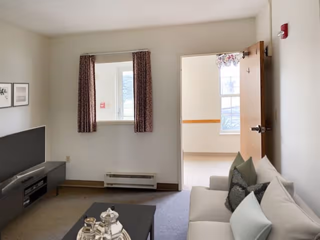 An assisted-living living room with a sofa and cushions, coffee table holding a tea set, TV stand, a curtained window, and an open door to a bright adjoining room.