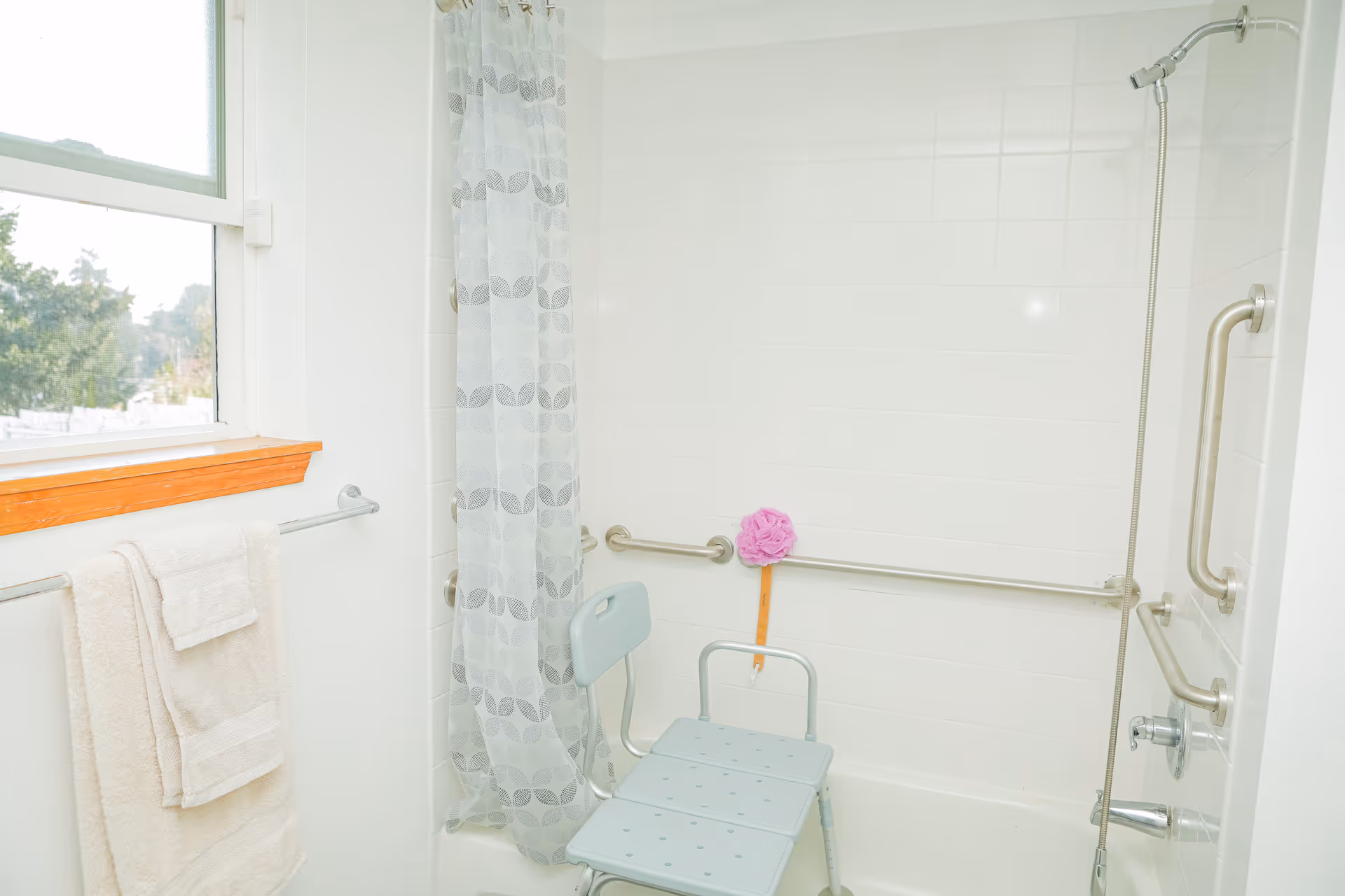 A bathroom with a white tiled shower area featuring a shower chair, grab bars, and a handheld showerhead. A window with a wooden frame is on the left side, and a towel rack with two white towels is mounted below the window. A translucent shower curtain with a circular pattern hangs inside the tub.