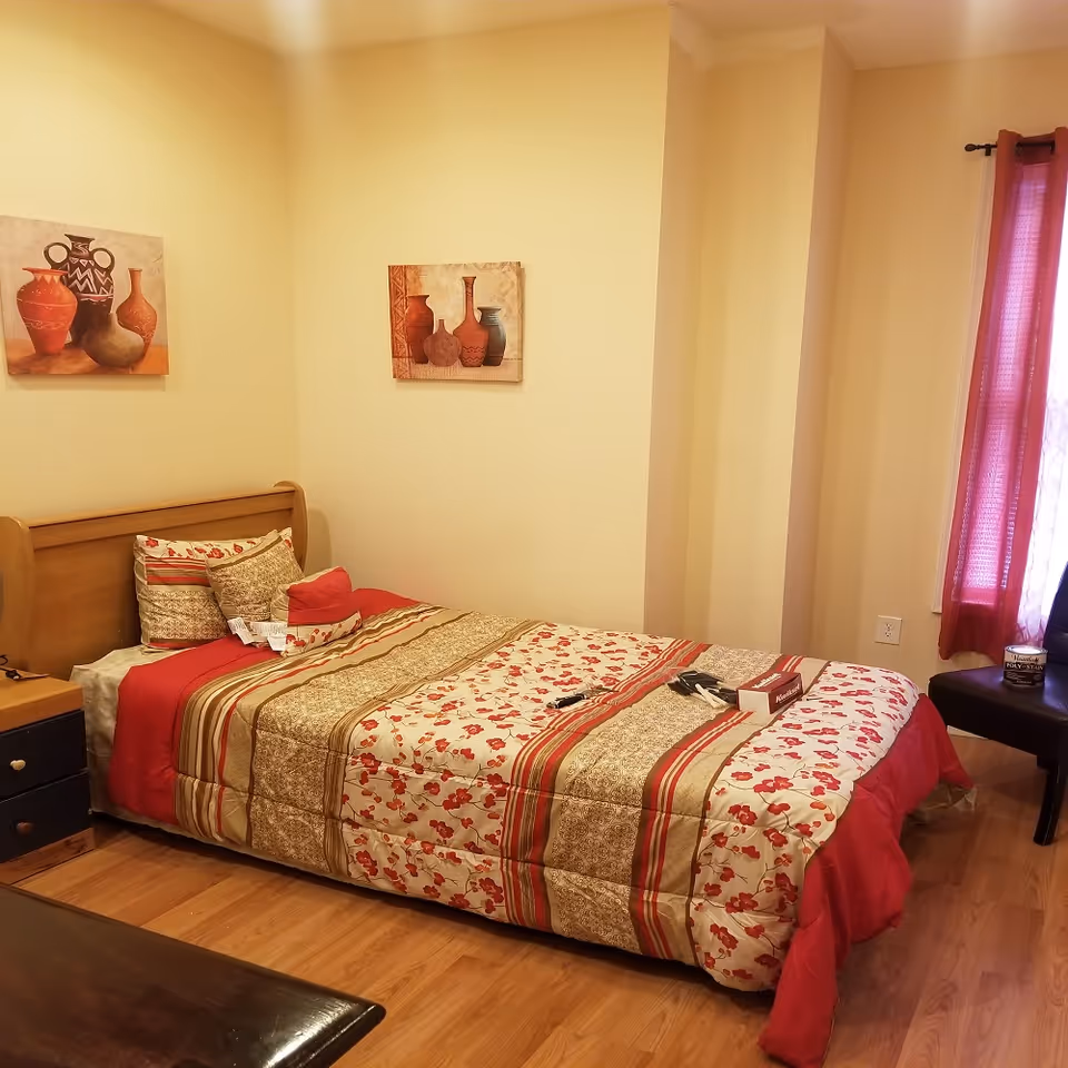 A bedroom with a single bed covered in a floral and striped comforter in shades of red, beige, and brown. There are multiple pillows on the bed. The room has light yellow walls with two paintings of vases hanging above the bed. A small nightstand with drawers is next to the bed, and a window with red curtains is on the right side. A black chair and a can of wood stain are near the window. The floor is wooden.