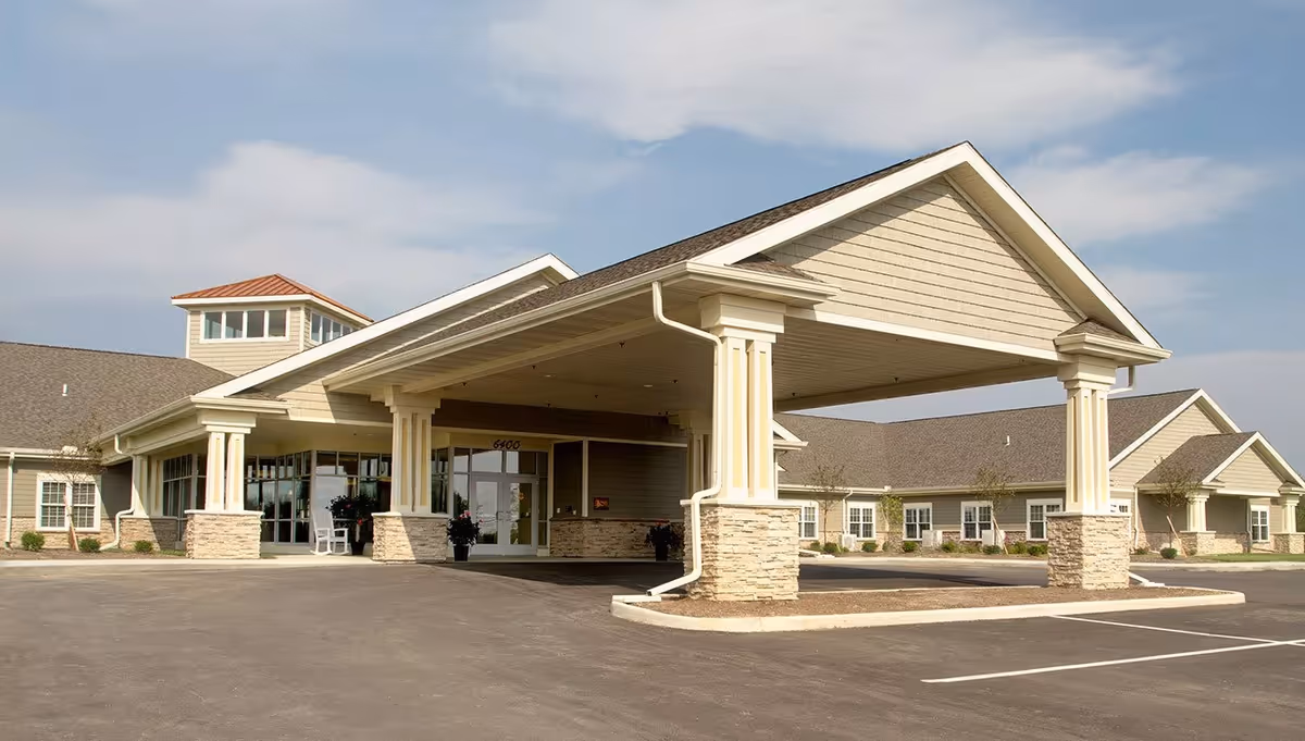 Exterior view of Randall Residence of Tipp City, showing a large covered entrance with stone pillars and beige siding, surrounded by a paved driveway and parking area under a partly cloudy sky.