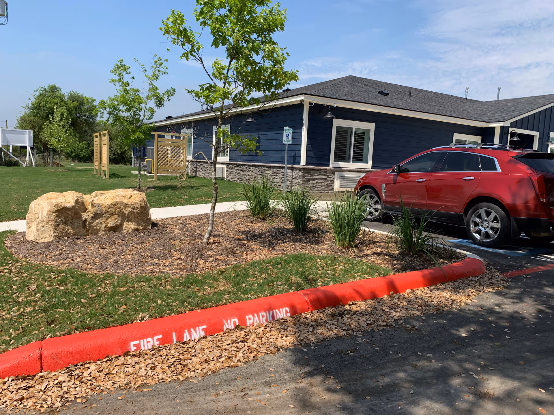 Exterior view of a single-story building with blue siding and stone accents, a red SUV parked in a parking space, a small landscaped area with young trees and plants, and a red curb painted with 'FIRE LANE NO PARKING'.