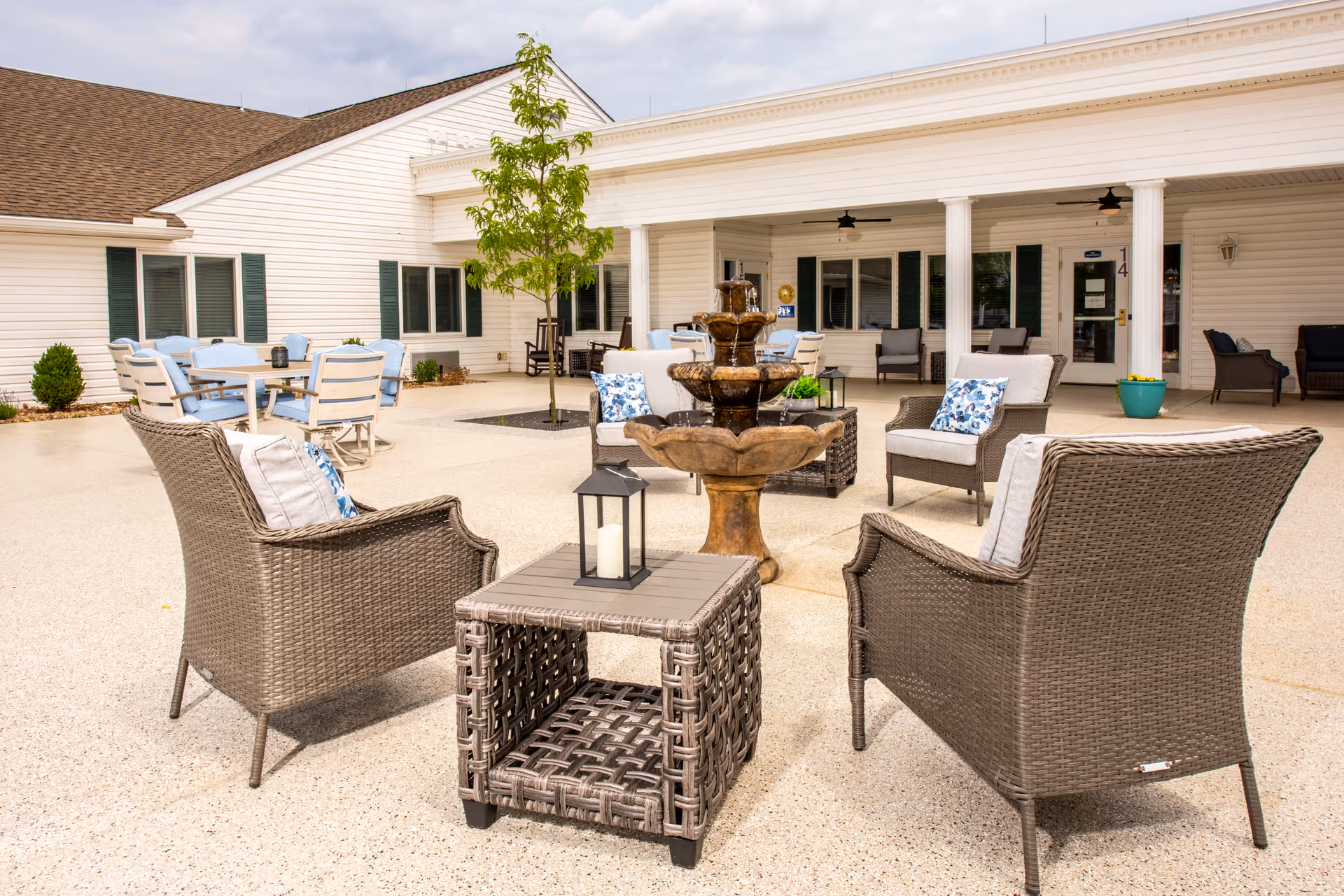 Outdoor patio area at Providence Place Senior Living of Dover featuring wicker chairs with cushions arranged around a central water fountain. There are additional tables and chairs in the background near the building with white siding and green shutters. A small tree is planted in the patio area.