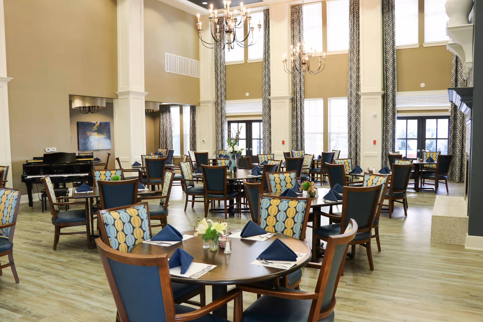 A spacious dining room with multiple round wooden tables and chairs featuring patterned upholstery and blue cushions. Each table is set with blue folded napkins, silverware, and small floral centerpieces. The room has high ceilings with large windows covered by patterned curtains, and two chandeliers hanging from the ceiling. A black grand piano is visible in the background.