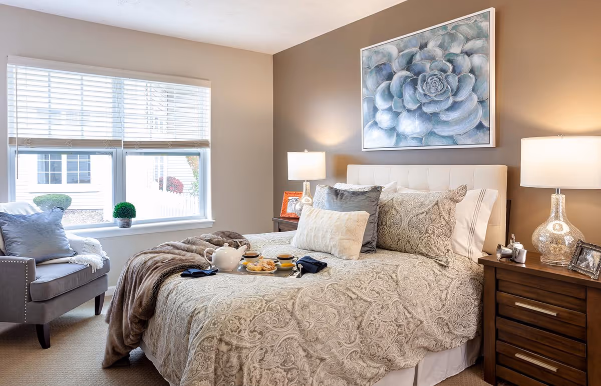 A cozy bedroom with a large bed covered in patterned bedding and multiple pillows. On the bed is a tray with a teapot, cups, and pastries. There are two bedside tables with lamps and picture frames. A gray armchair with a pillow and blanket is placed near a window with white blinds. A large painting of a succulent hangs on the wall above the bed.