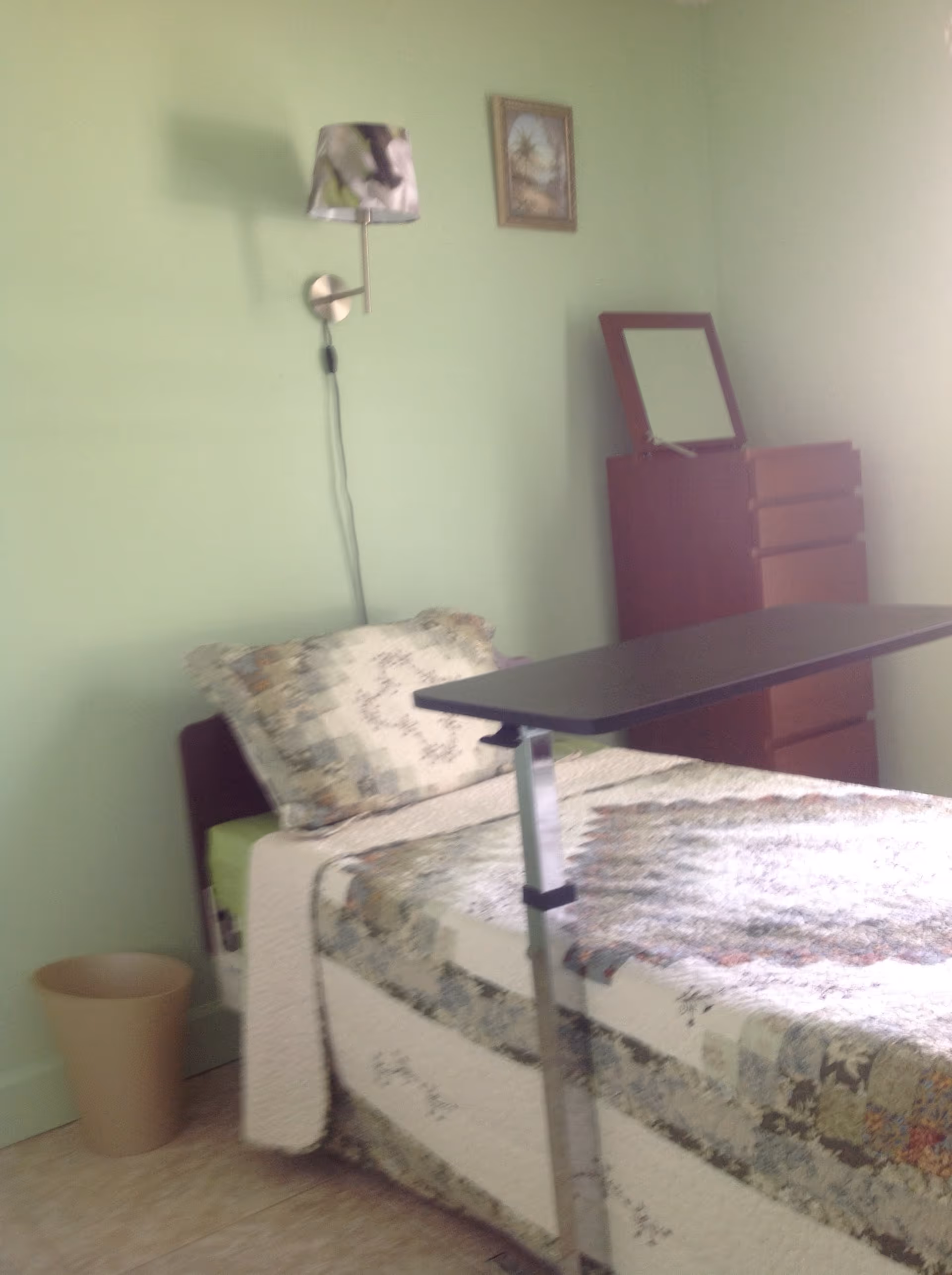 A small bedroom with a single bed covered in a patchwork quilt. There is a pillow matching the quilt, a wall-mounted lamp with a patterned shade, a wooden dresser with a mirror on top, and a beige wastebasket on the floor. The walls are painted light green.