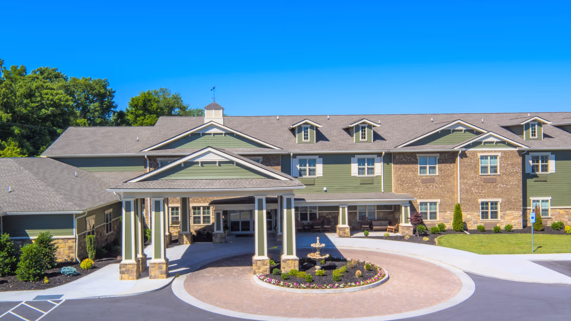 Front exterior view of Sycamore Springs Senior Living Community building with a covered entrance, circular driveway, landscaped flower bed with a small fountain, and clear blue sky.