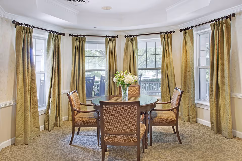 Bright dining area with a round wooden table and four upholstered chairs in front of large bay windows with green curtains and a vase of white flowers.