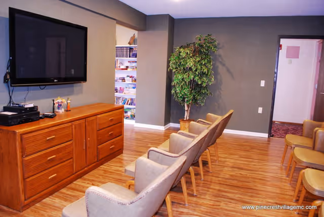 A common area with a row of beige cushioned chairs facing a wall-mounted flat screen TV above a wooden cabinet. The room has wood flooring, gray walls, and a potted plant in the corner. A doorway leads to another room with shelves filled with books and games.