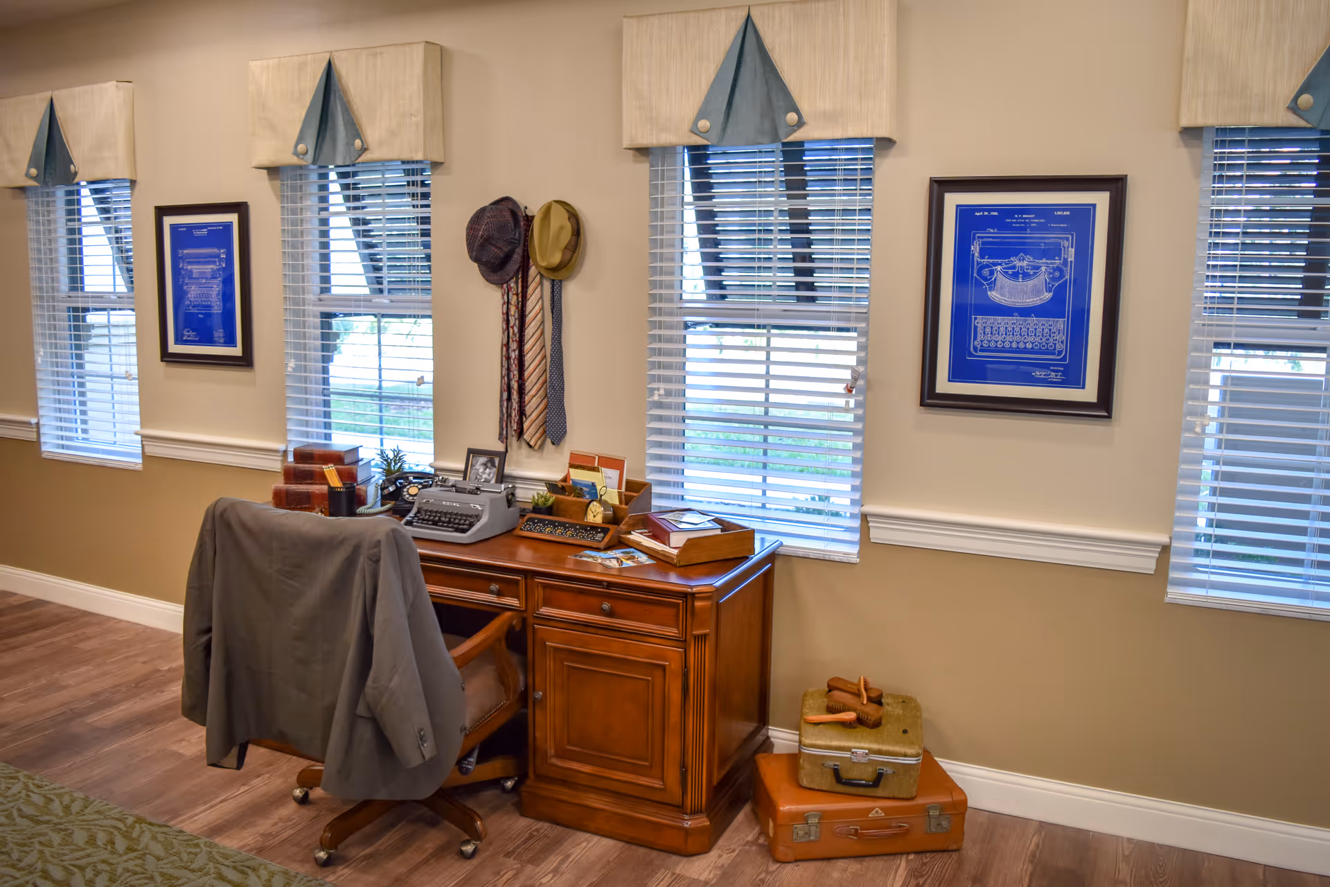 A cozy office corner in a senior living facility with a wooden desk and chair. The desk holds a vintage typewriter, decorative boxes, a small plant, and various office items. A gray jacket is draped over the chair. Three windows with blinds and beige valances let in natural light. Two framed blueprints of a typewriter hang on the wall. Two hats and three ties hang on hooks between the windows. On the floor next to the desk are two vintage suitcases with a pair of wooden shoe stretchers on top.
