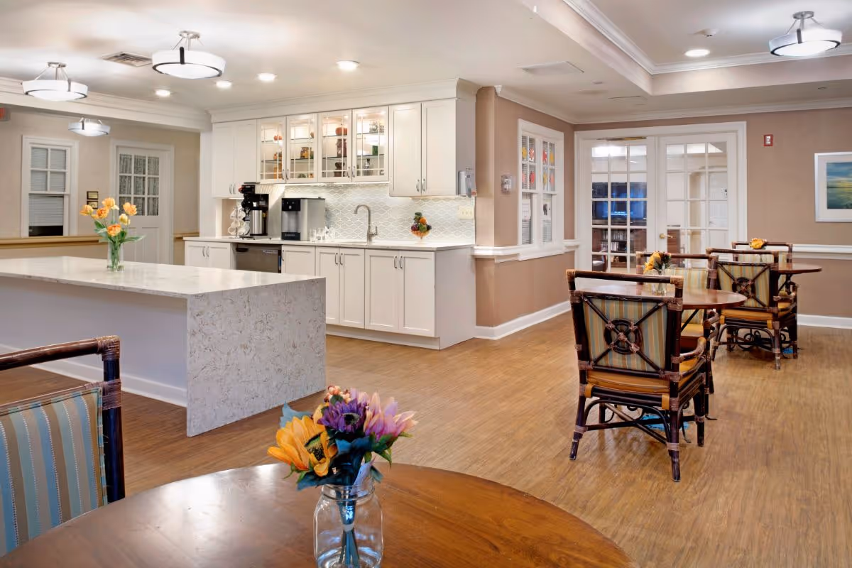 Bright communal dining area with a marble-topped kitchen island, white cabinets, and several tables with floral centerpieces.