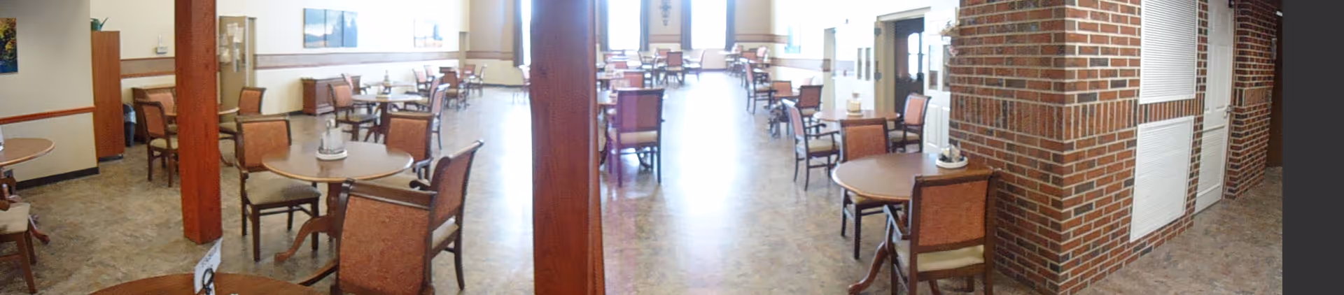 A spacious dining room with multiple round tables and chairs arranged neatly. The room has large windows allowing natural light to brighten the space. There are brick pillars and walls, and the floor is covered with a patterned tile. The room appears clean and well-organized, suitable for communal dining.
