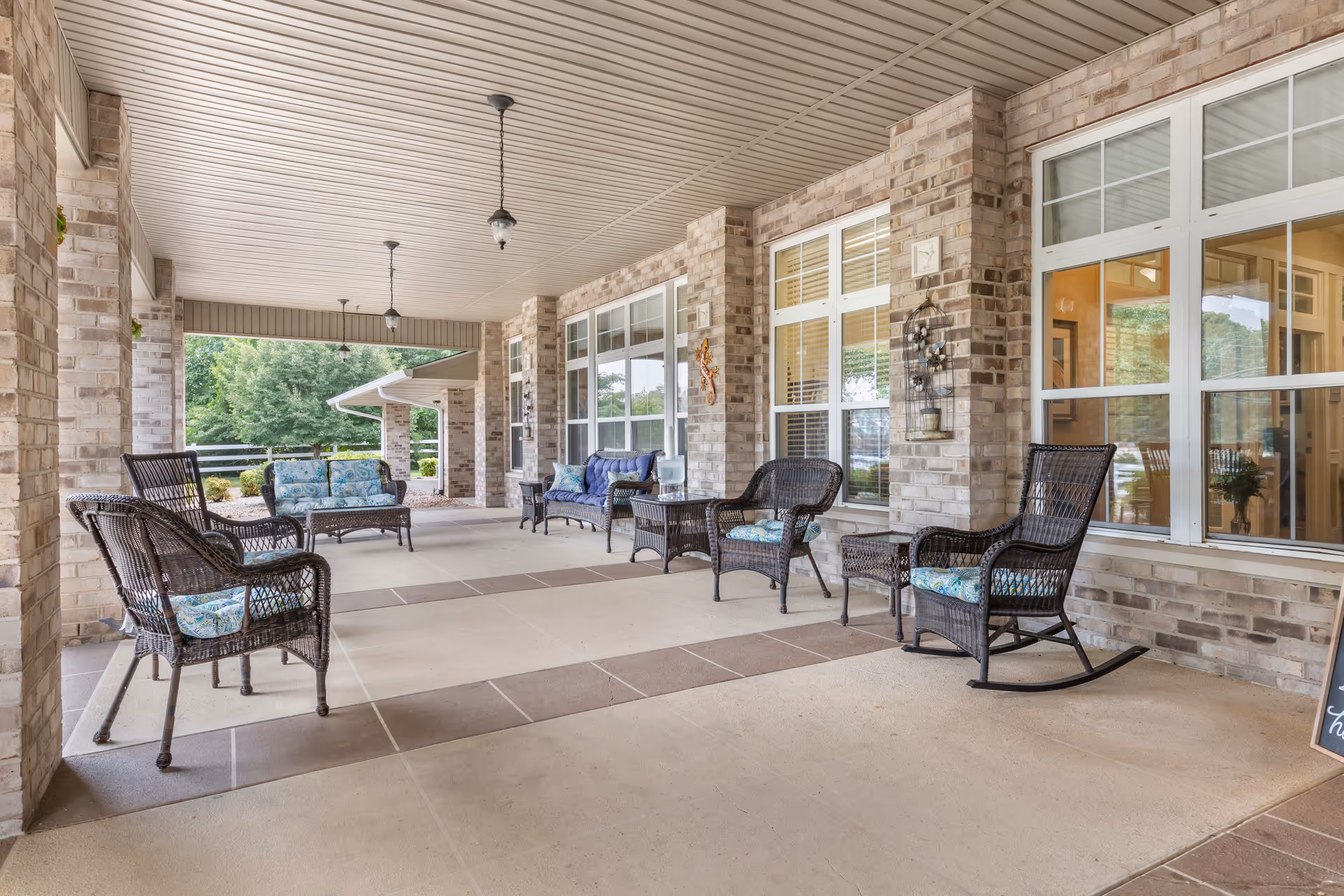 Covered outdoor patio area with wicker chairs and sofas featuring blue patterned cushions, brick walls, large windows, and hanging light fixtures. The patio overlooks a green garden area with trees and shrubs.