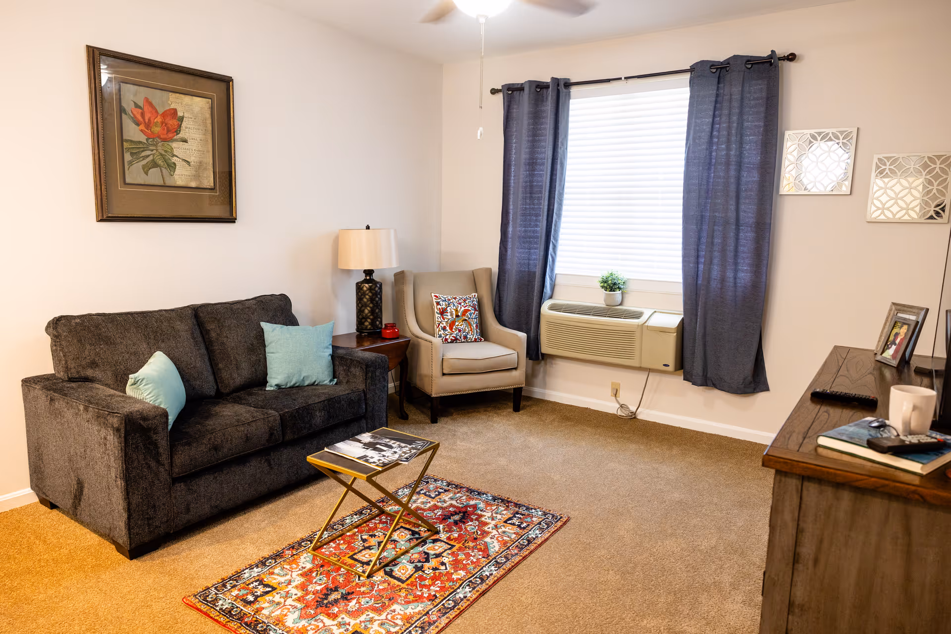 A cozy living room with a dark gray sofa adorned with light blue pillows, a beige armchair with a colorful patterned cushion, a small wooden side table with a lamp, and a small gold and glass coffee table on a vibrant red patterned rug. The room has beige carpet, a window with blue curtains and white blinds, an air conditioning unit below the window, and a wooden dresser with books, a remote control, a coffee mug, and framed photos on top. A framed floral artwork hangs on the wall above the sofa.