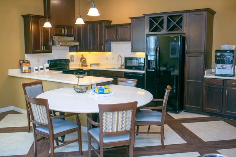 A modern kitchen area with dark wooden cabinets, a black refrigerator, microwave, stove, and coffee machine. There is a round white table with four wooden chairs around it, and some items including a box of Glad ClingWrap and a book on the table. The floor has a patterned design with wood and tile.