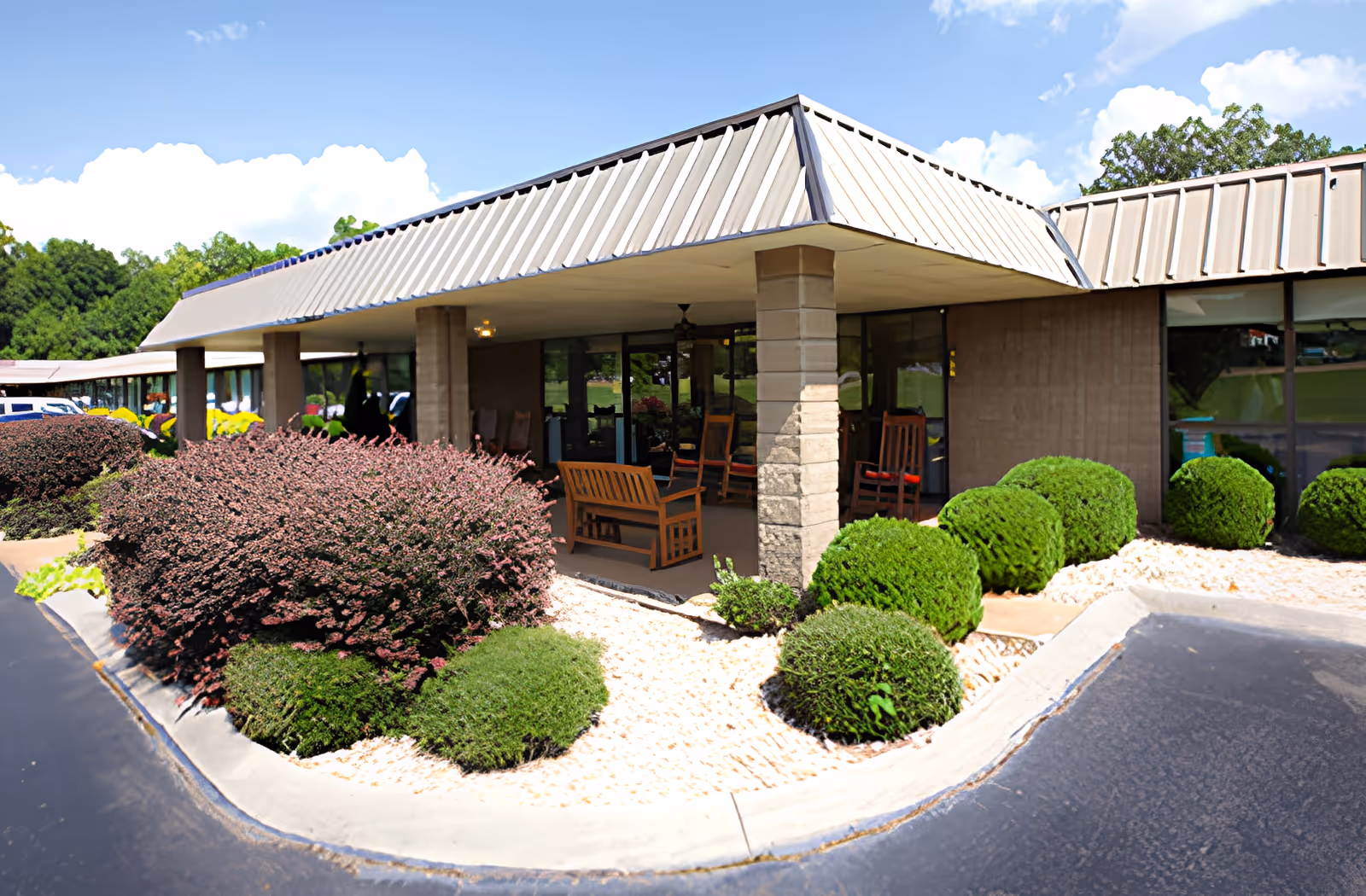 Exterior view of Ahc West Tennessee Transitional Care facility showing a covered patio area with wooden rocking chairs and benches. The building has large windows and a metal roof. The surrounding landscape includes neatly trimmed bushes and small white rocks along the edge of the pavement under a bright blue sky with some clouds.