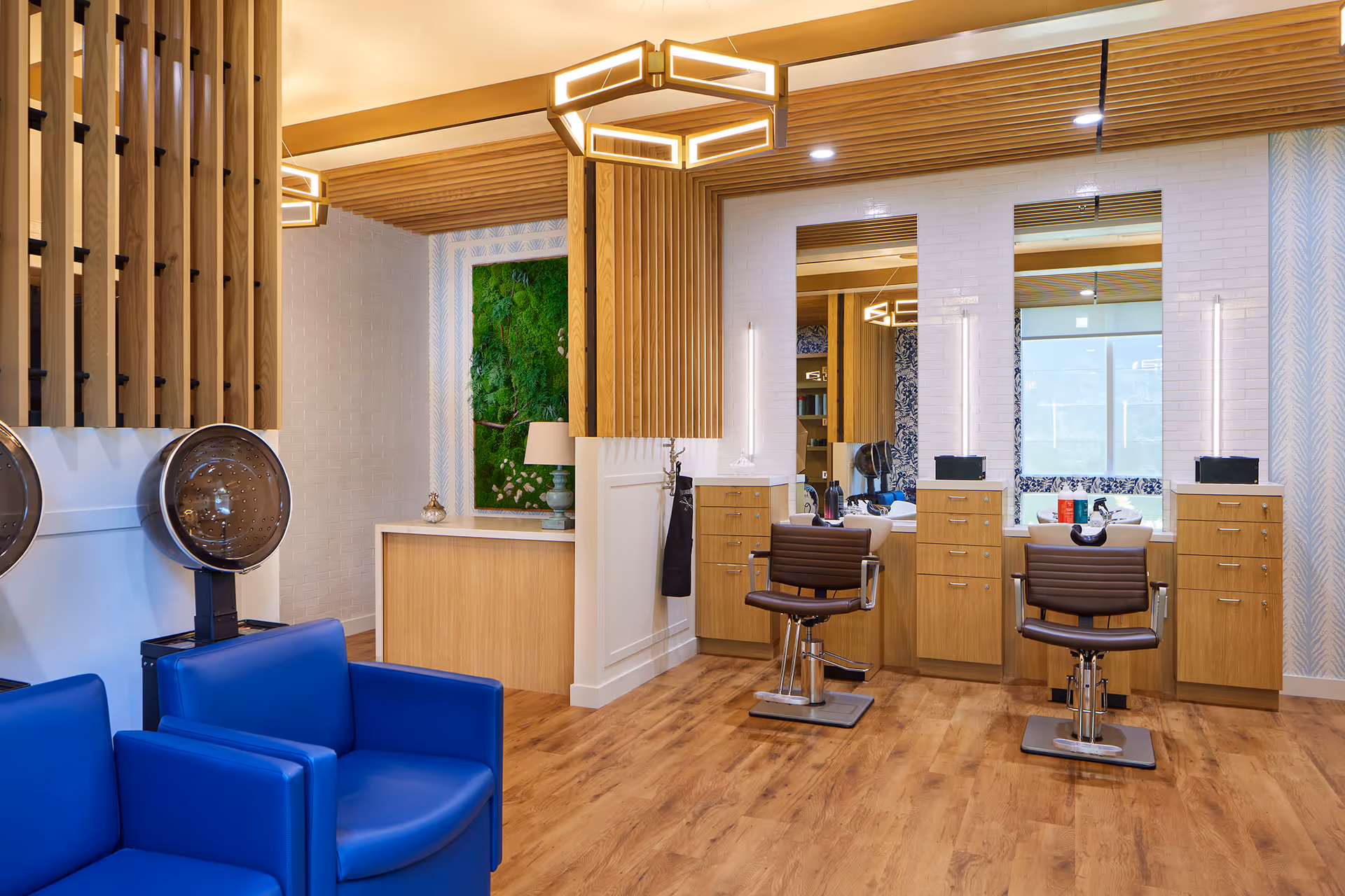 Interior view of a modern salon area in a senior living facility featuring two brown salon chairs in front of mirrors with wooden cabinetry, blue armchairs, hair dryers mounted on the wall, wooden ceiling accents, and a green plant wall decoration.