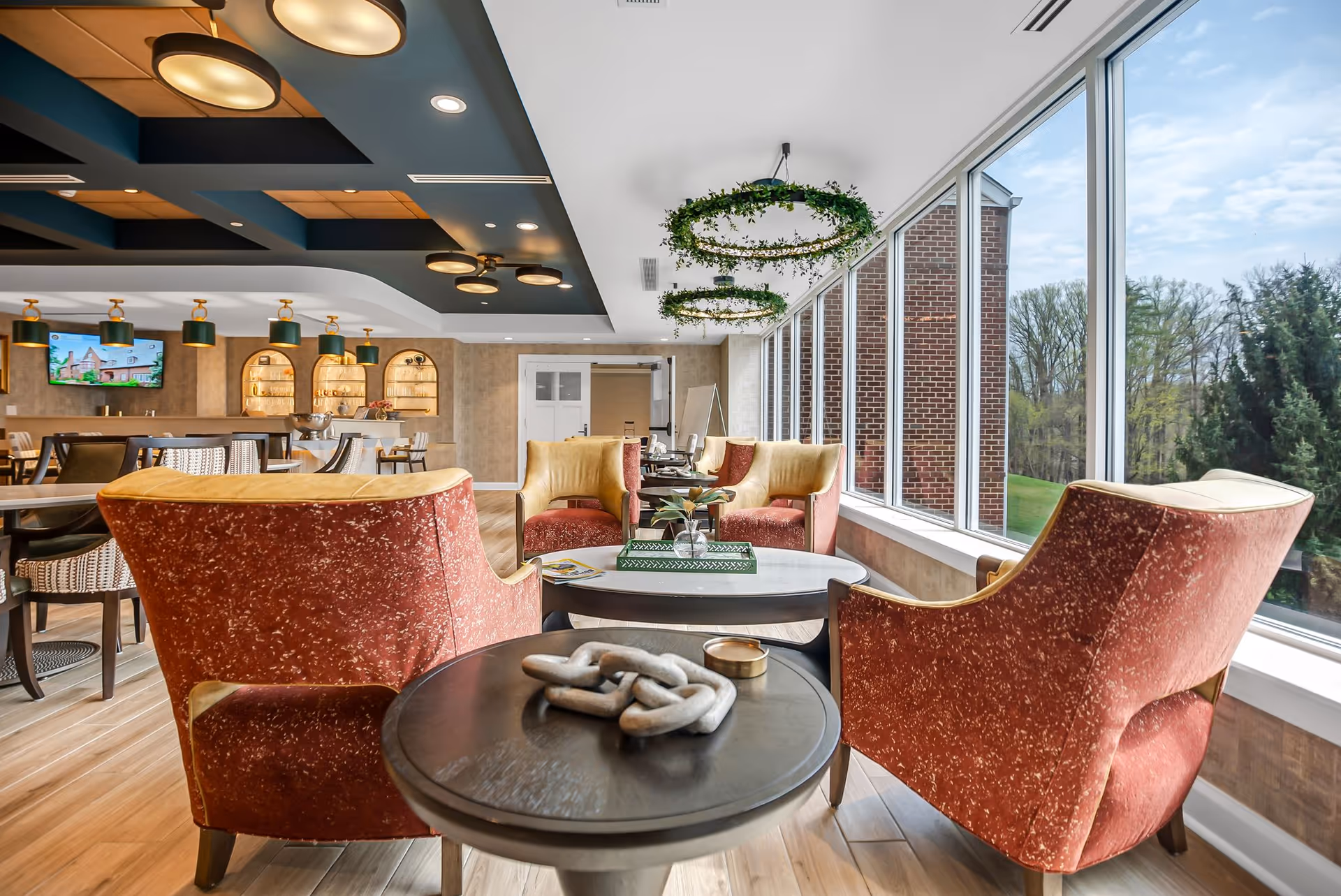 A bright and spacious lounge area with large windows offering a view of trees outside. The room features comfortable red and yellow armchairs arranged around a coffee table with decorative items. The ceiling has modern light fixtures and hanging green wreaths. In the background, there is a dining area with tables, chairs, and a bar with shelves and pendant lights.