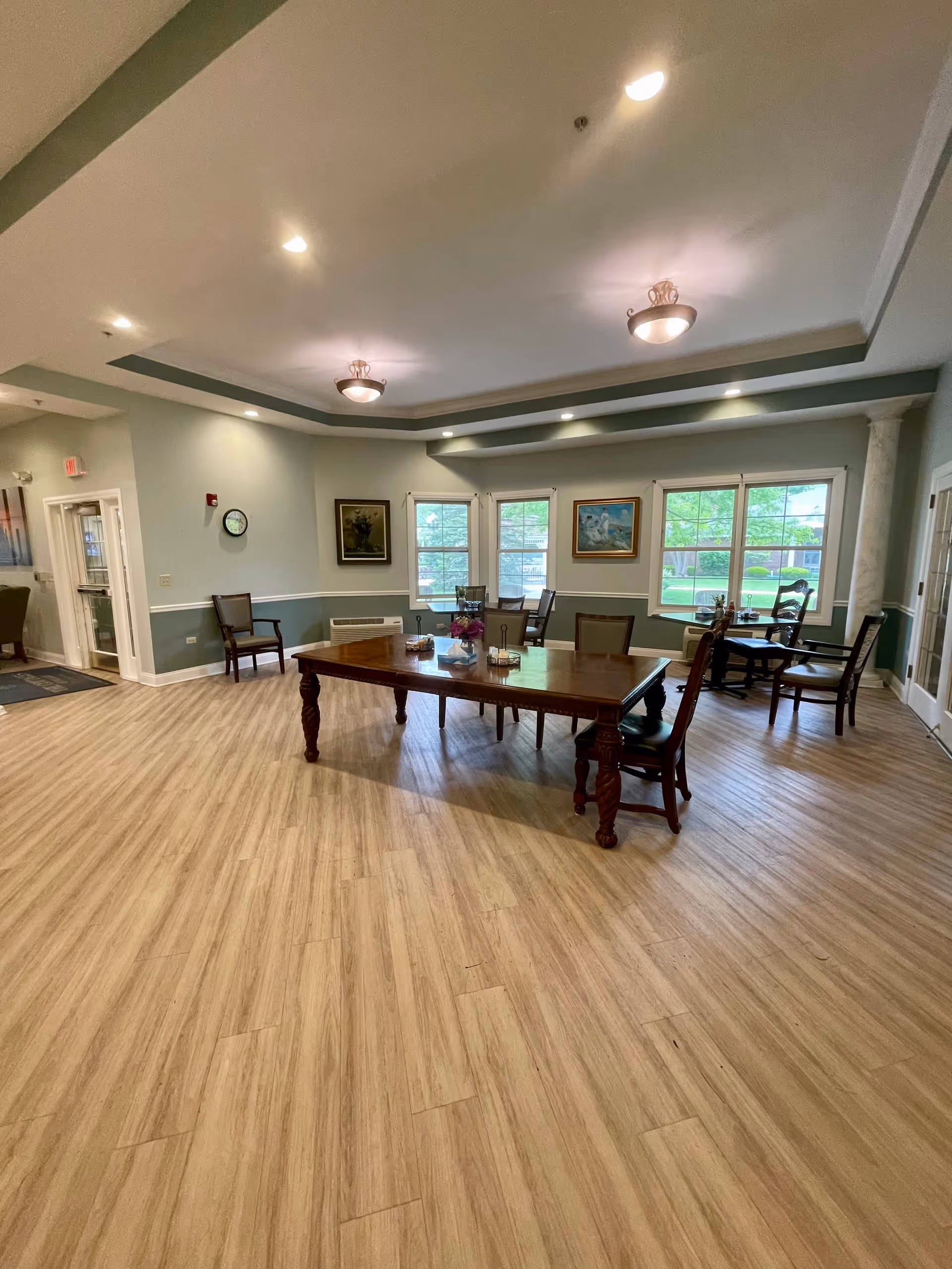 A spacious common area with wooden flooring and a large wooden table surrounded by chairs in the center. The room has light green walls with white trim, several windows letting in natural light, framed paintings on the walls, and ceiling lights. Additional chairs and small tables are placed near the windows.