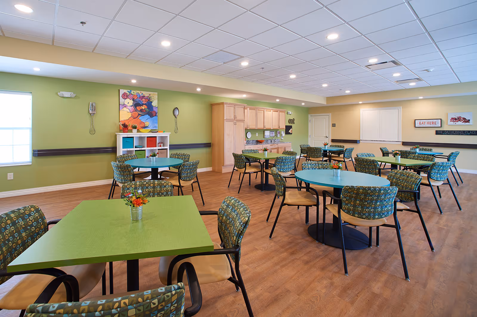 A bright and spacious dining room with green and blue tables surrounded by patterned chairs. The room has light green and beige walls, wooden flooring, and a white ceiling with recessed lighting. There are decorative wall hangings including a colorful painting, oversized fork and spoon, and signs that say 'EAT HERE' and 'THE GATHERING PLACE'. A white shelving unit with colorful bins and a wooden cabinet with a countertop are also visible.