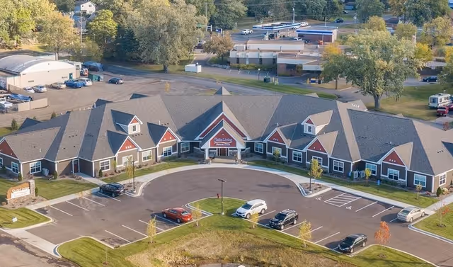 Aerial view of Suite Living Senior Care of West St. Paul building with a large parking lot in front, surrounded by trees and greenery.