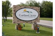 A brick roadside sign reading "Cortland Acres" set on a grassy lawn with small planters and trees in the background.