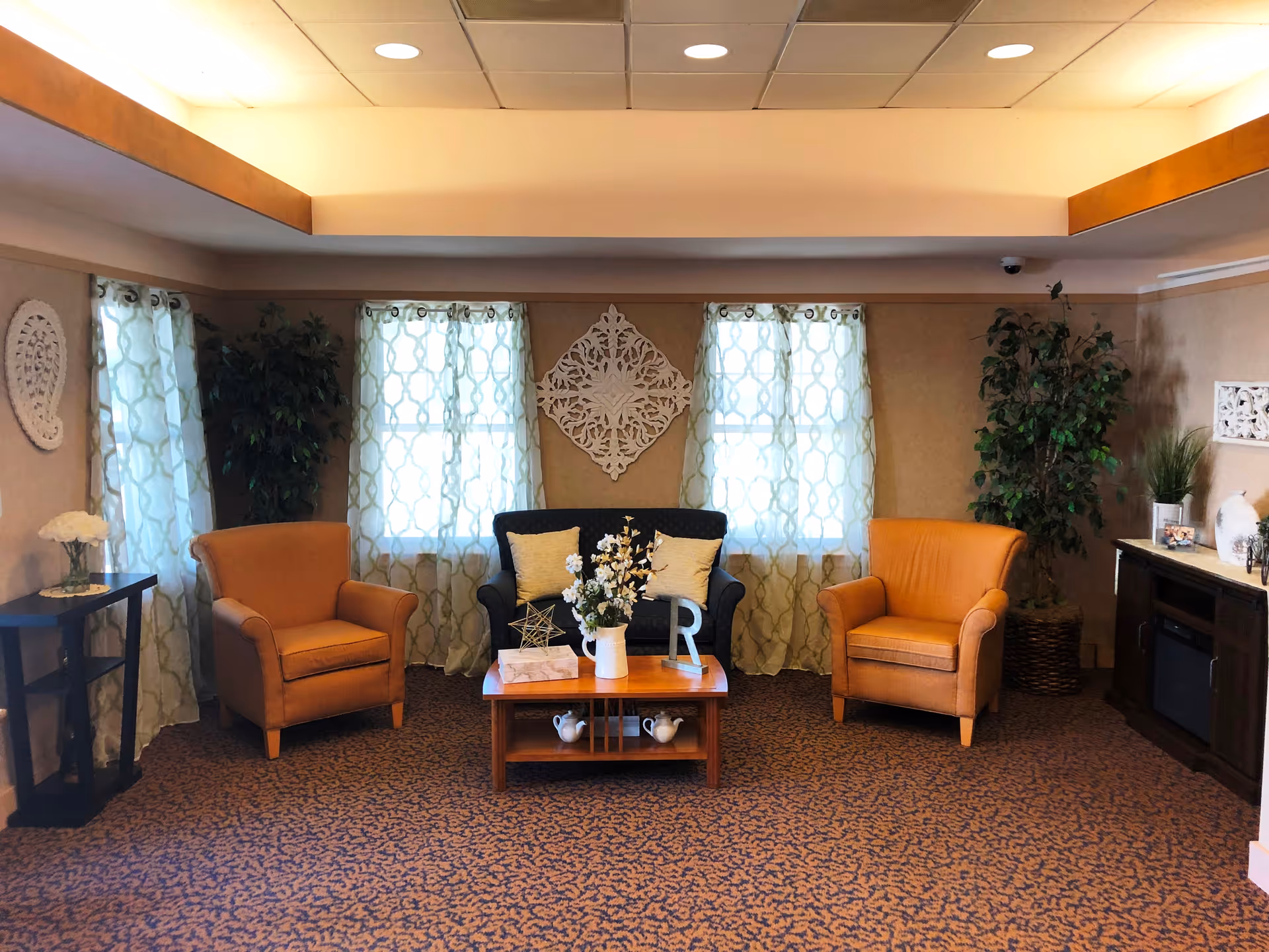 A cozy living room area with two orange armchairs and a dark blue loveseat arranged around a wooden coffee table decorated with a vase of flowers and decorative items. The room has patterned curtains on three windows, beige walls with decorative wall hangings, a patterned carpet, and green plants in the corners. There is a small side table with a flower vase on the left and a wooden cabinet with decorative items on the right.