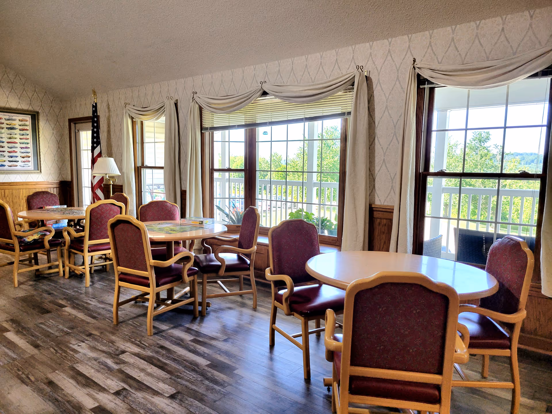 A bright dining area with round wooden tables and maroon cushioned chairs. Large windows with cream-colored curtains allow natural light to fill the room, and an American flag stands in the corner next to a floor lamp. The floor has a wood-like finish, and there is a framed picture on the wall.