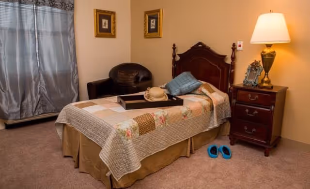A cozy bedroom with a single bed covered in a patchwork quilt, a wooden headboard, and a tray with a hat and pillow on the bed. Next to the bed is a wooden nightstand with a lamp, a picture frame, and a small decorative item. A pair of blue slippers are on the carpeted floor beside the nightstand. In the corner, there is a black armchair and a window with gray curtains.