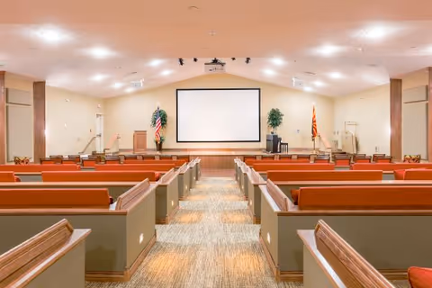 Interior view of a chapel-style meeting room with rows of pews facing a stage and large projection screen.