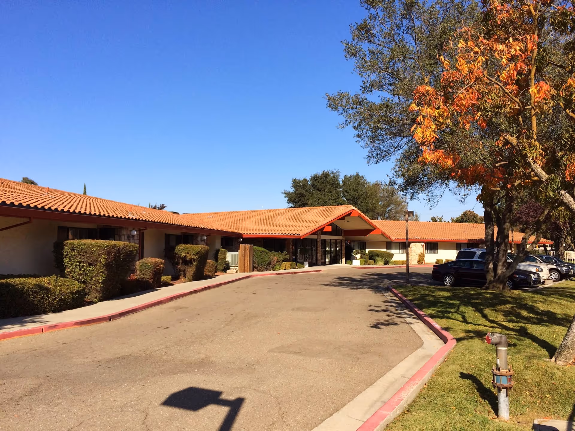 Exterior view of Riverwood Healthcare Center showing a single-story building with a red-tiled roof, trimmed bushes along the walkway, a driveway leading to the main entrance, several parked cars, and a tree with autumn-colored leaves on the right side under a clear blue sky.