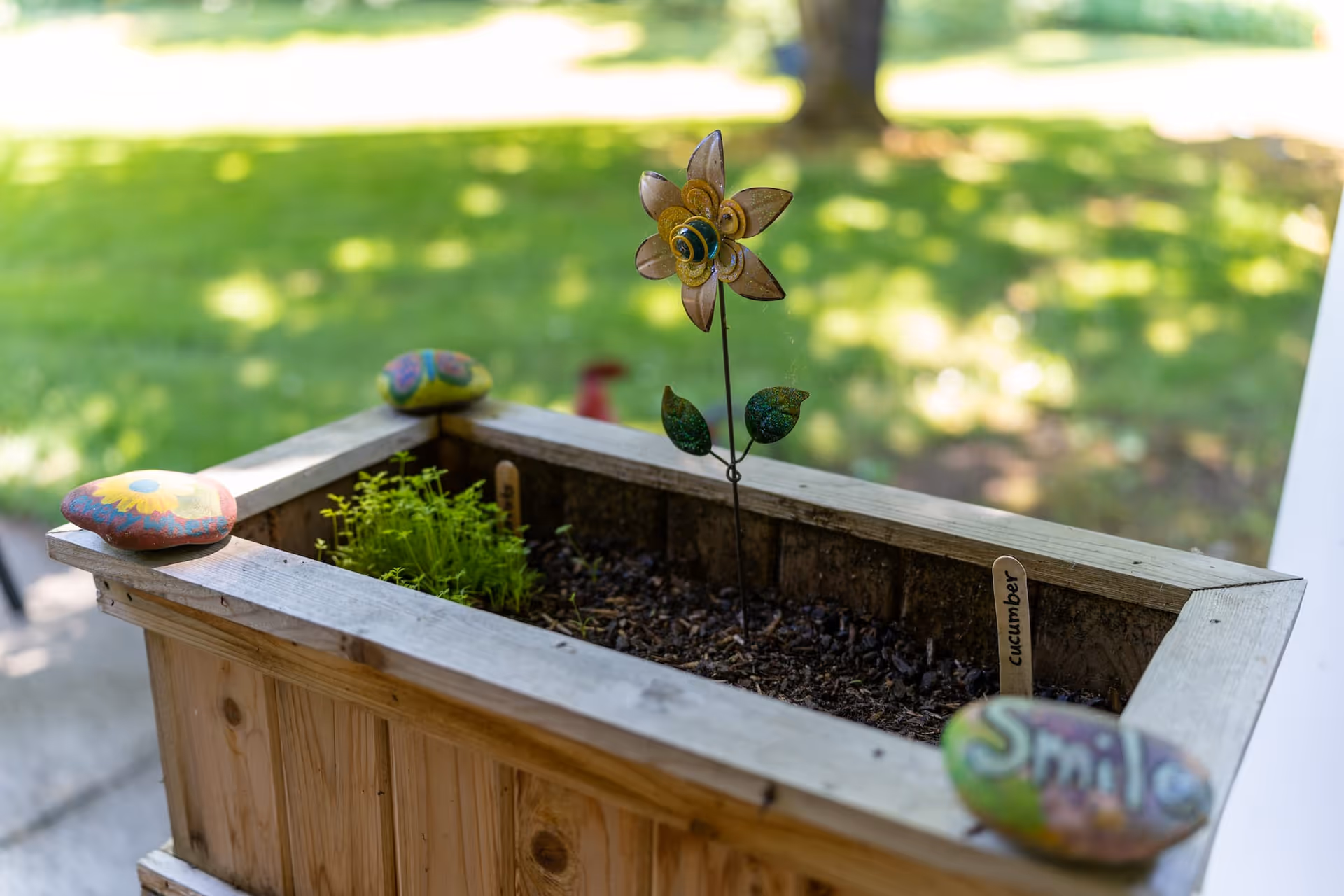 Wooden planter box on a porch with a decorative metal flower stake, small herbs, and painted rocks set against a blurred grassy yard.