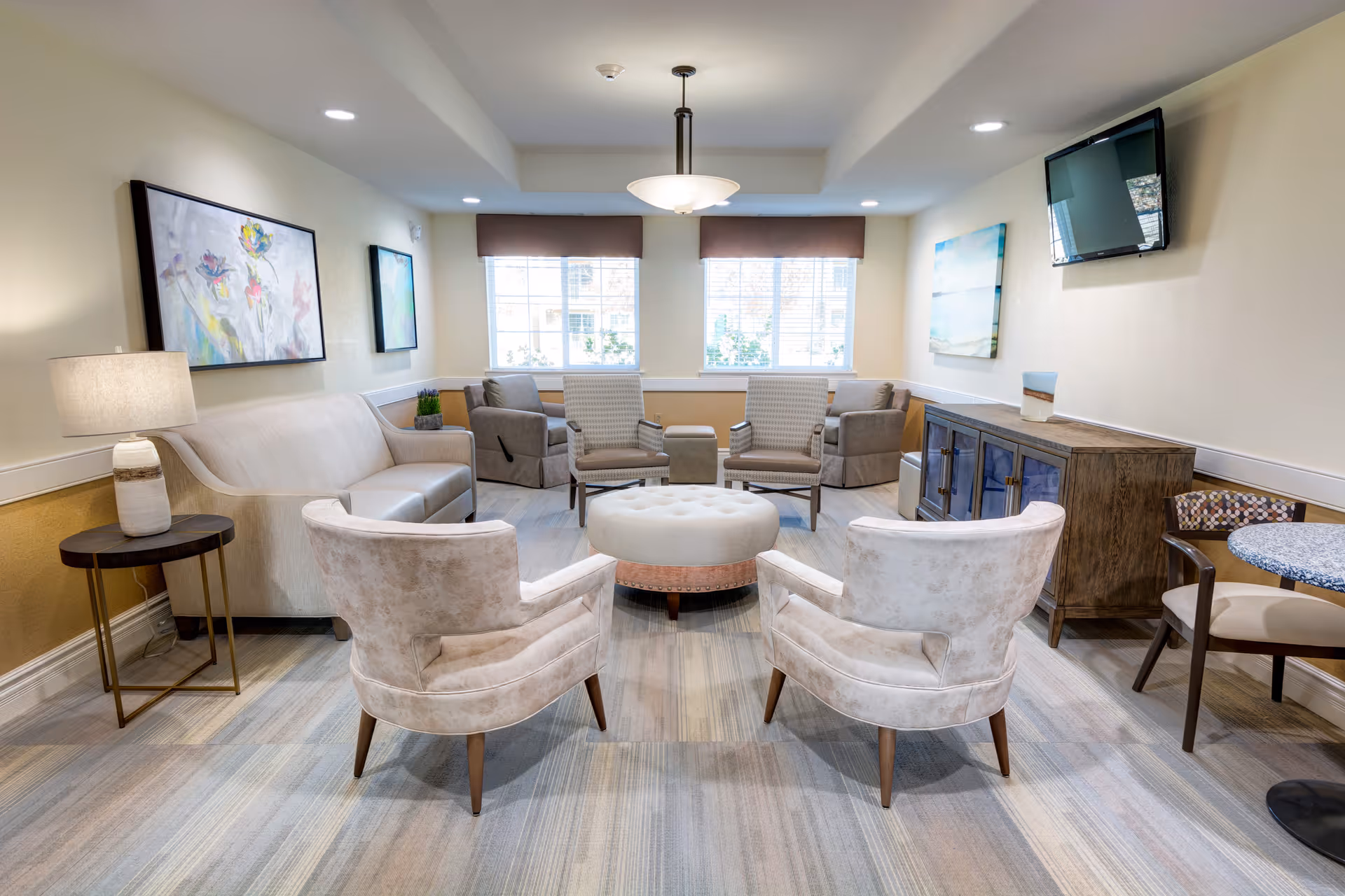 A cozy senior living common area with a beige sofa, two beige armchairs, two patterned chairs, a round ottoman in the center, a wooden sideboard with a small painting on top, a wall-mounted TV, and two large windows with brown valances letting in natural light. The room is decorated with framed artwork and has a soft, neutral color palette.