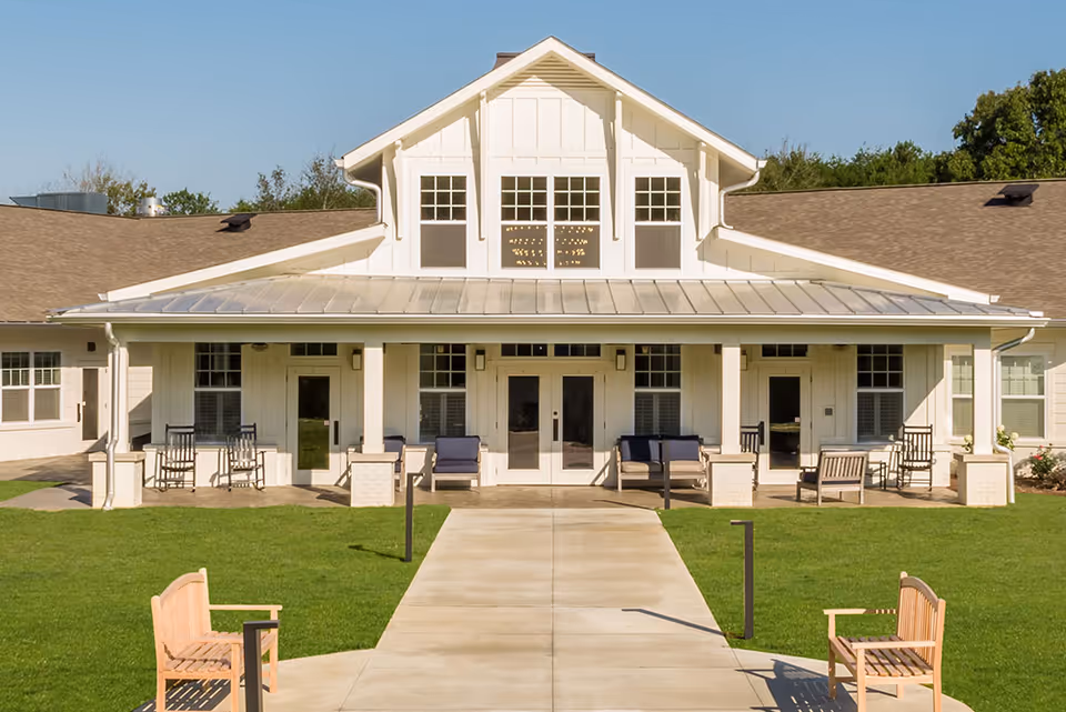 Front exterior view of a single-story senior living facility building with a covered porch featuring rocking chairs and benches, a concrete walkway leading to the entrance, and a well-maintained green lawn under a clear blue sky.