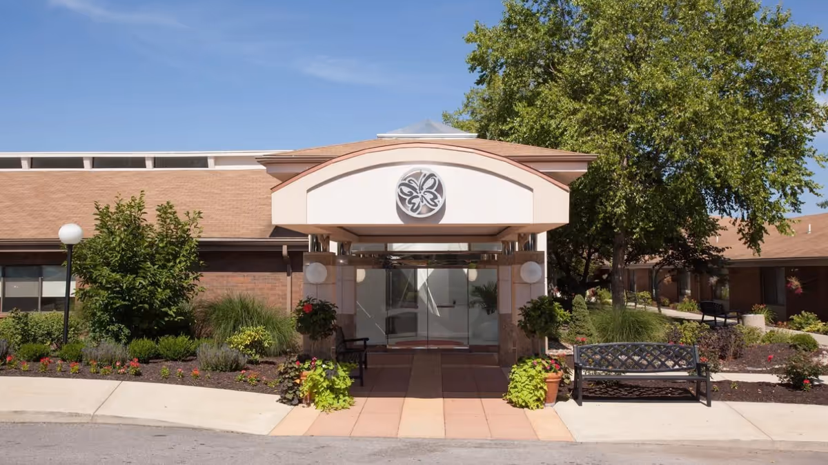 Front entrance of a senior living facility with a covered awning, glass doors, landscaped flowerbeds and benches.