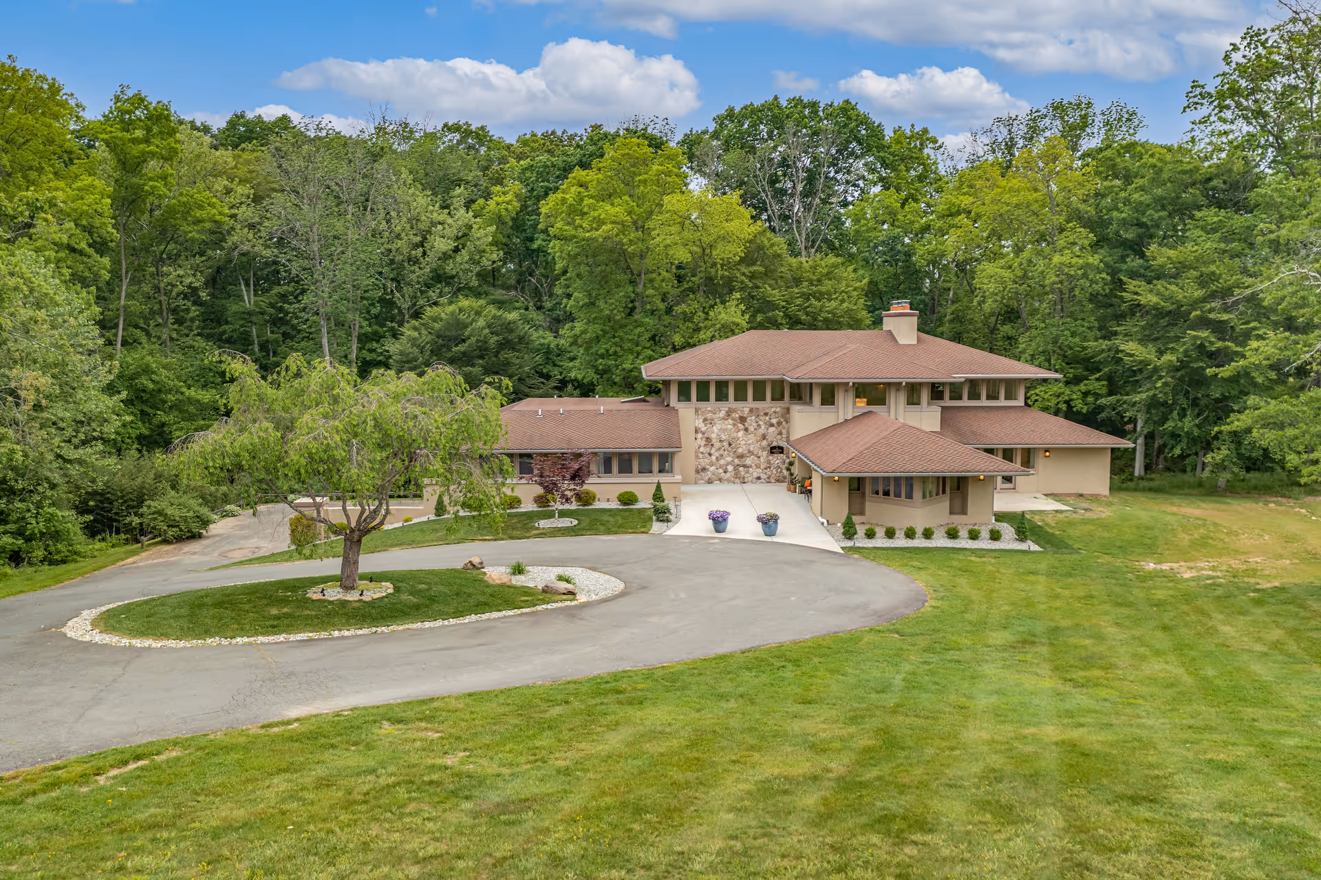 A large, two-story house with a brown roof and beige exterior surrounded by lush green trees and grass. There is a circular driveway with a tree in the center and two flower pots near the entrance. The sky is partly cloudy.