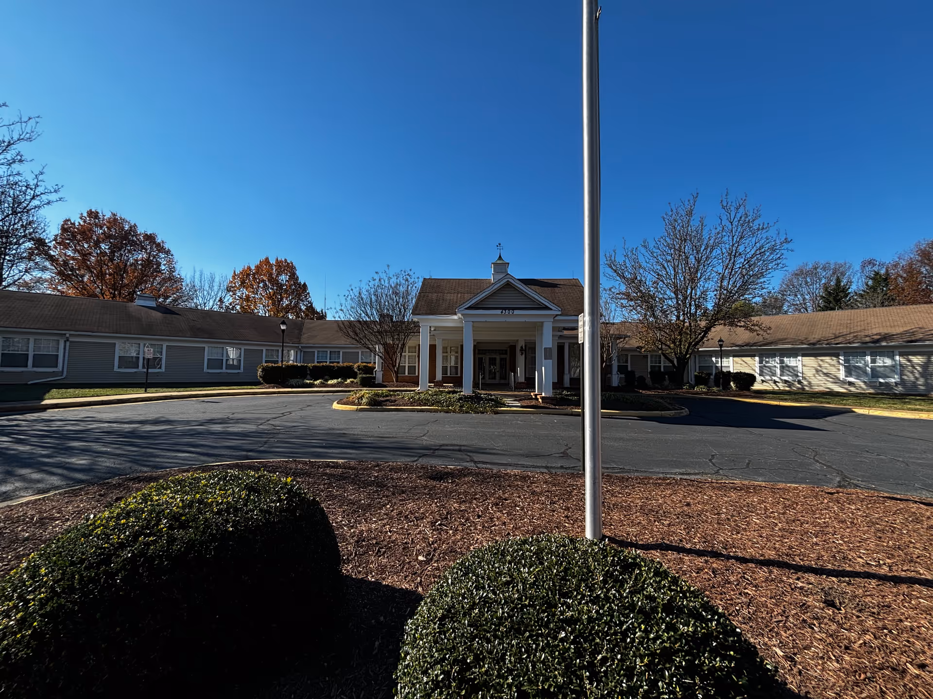 Front entrance of a single-story senior living facility with a portico, circular driveway, and landscaped shrubs under a clear blue sky.