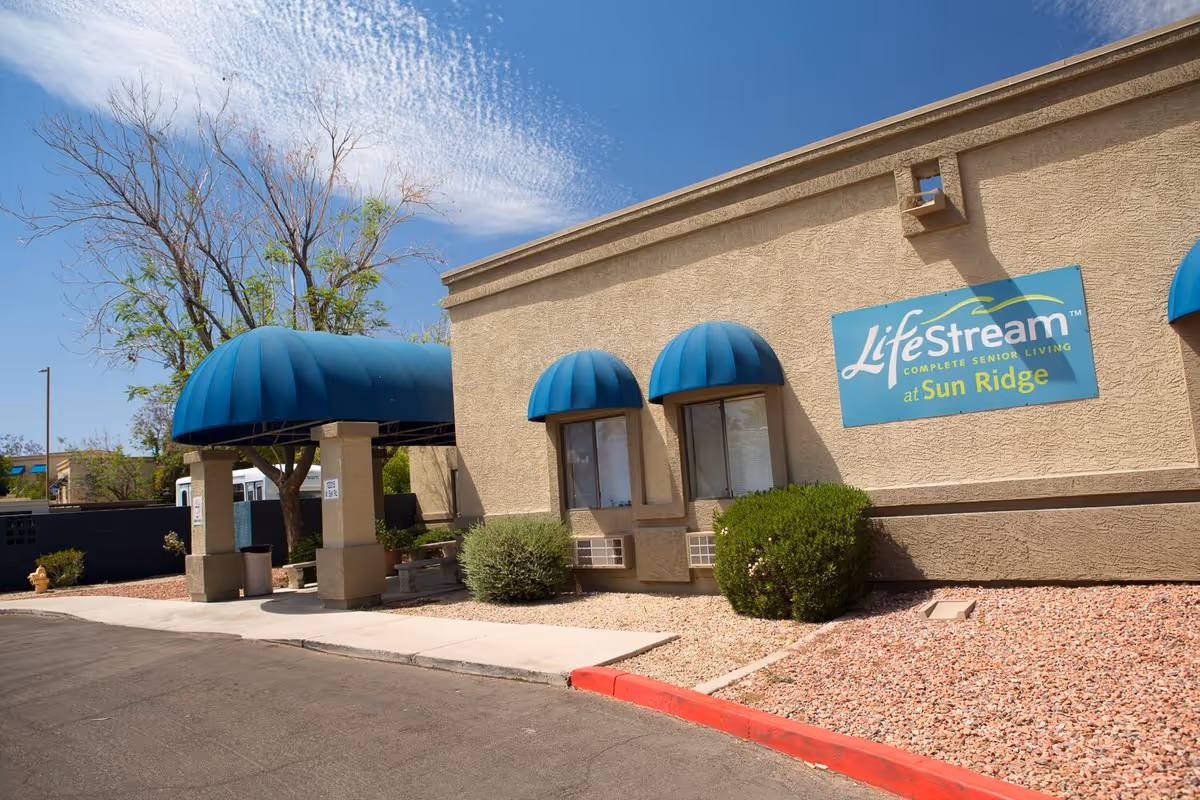 Front exterior of LifeStream Complete Senior Living at Sun Ridge showing blue entry awnings and the facility sign on a stucco wall.
