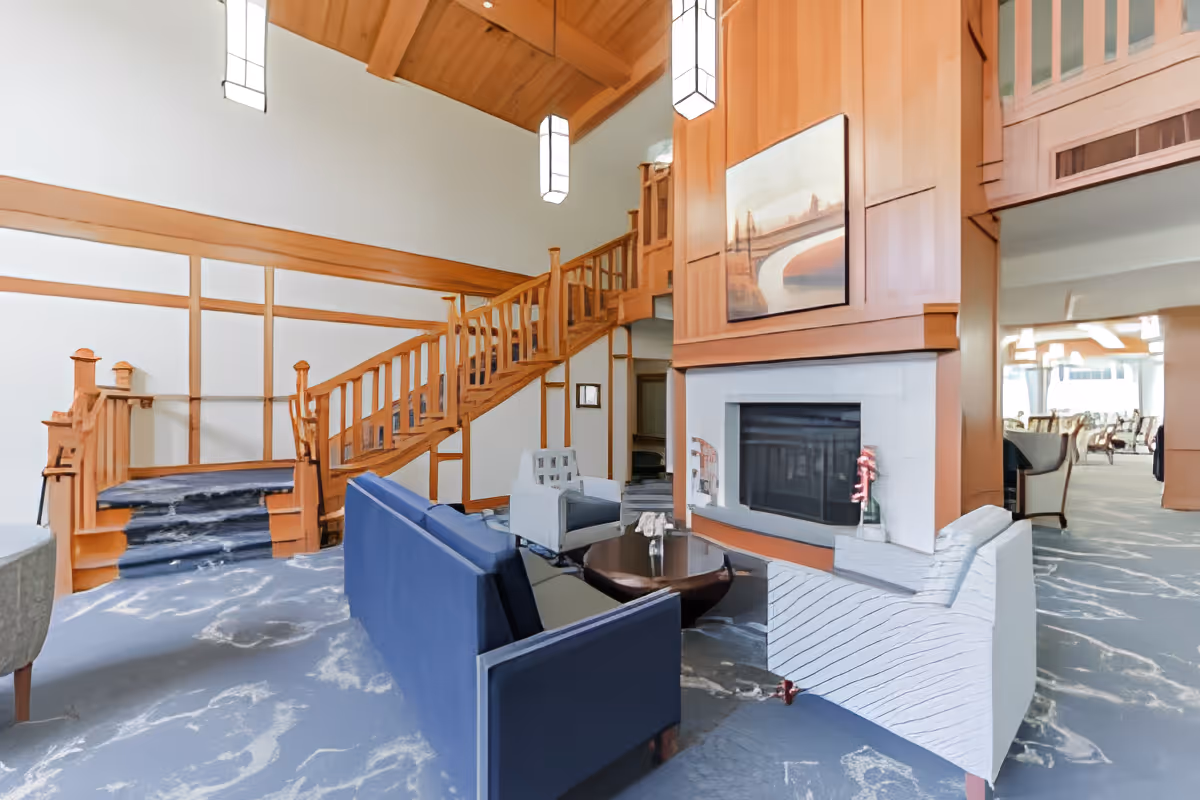 A spacious living room area with a high wooden ceiling and hanging pendant lights. The room features a wooden staircase with railings, a fireplace with a painting above it, and seating including a blue sofa and white armchairs arranged around a round coffee table. In the background, there is a dining area with multiple chairs and tables.